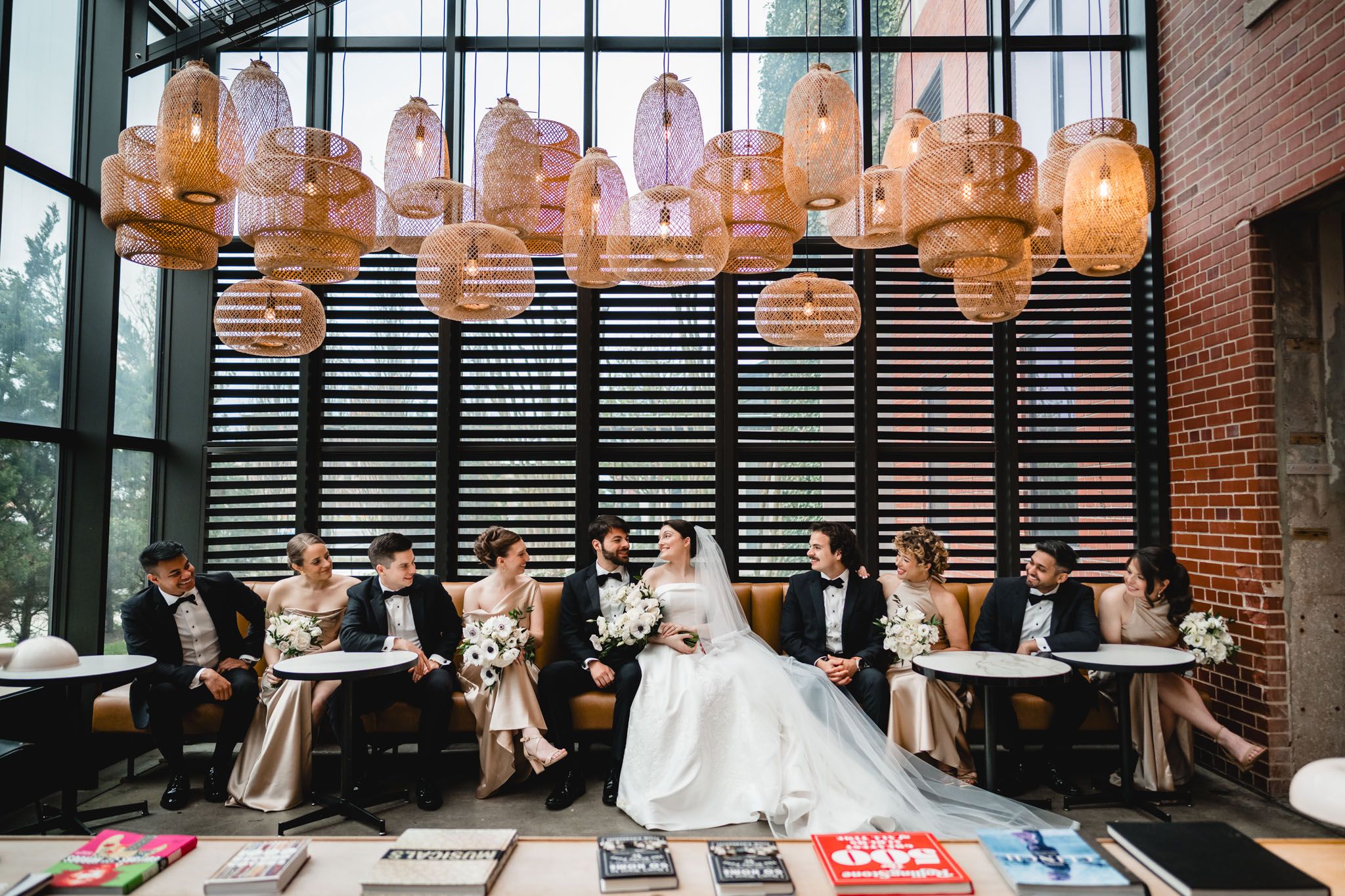 Wedding party sits together on lounge seating beneath decorative pendant lights in a modern venue space.
