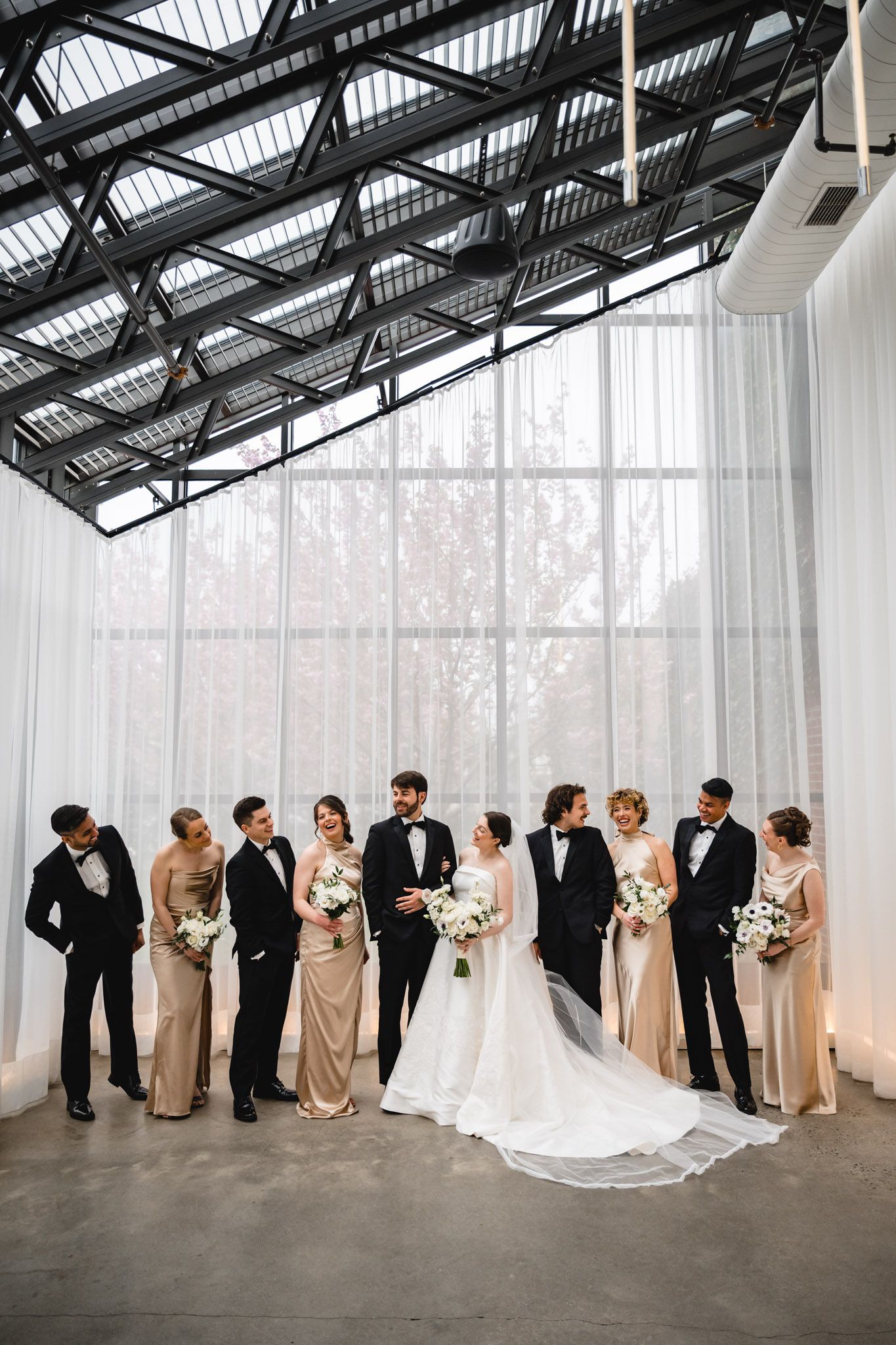 Wedding party in black tuxedos and champagne gowns pose with bride and groom in modern glass venue.