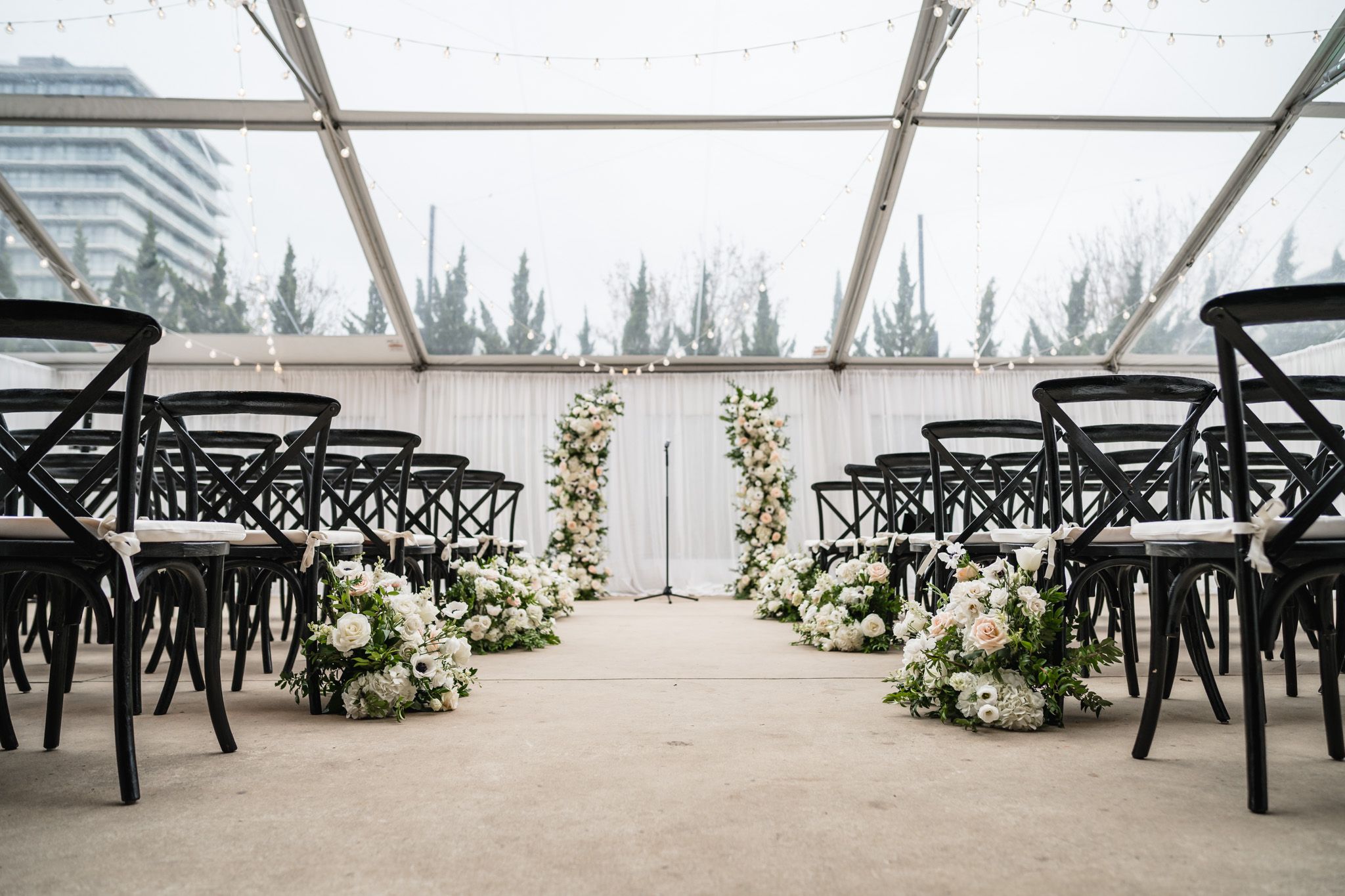 Elegant wedding ceremony aisle with white floral arrangements and black cross-back chairs in clear tent.