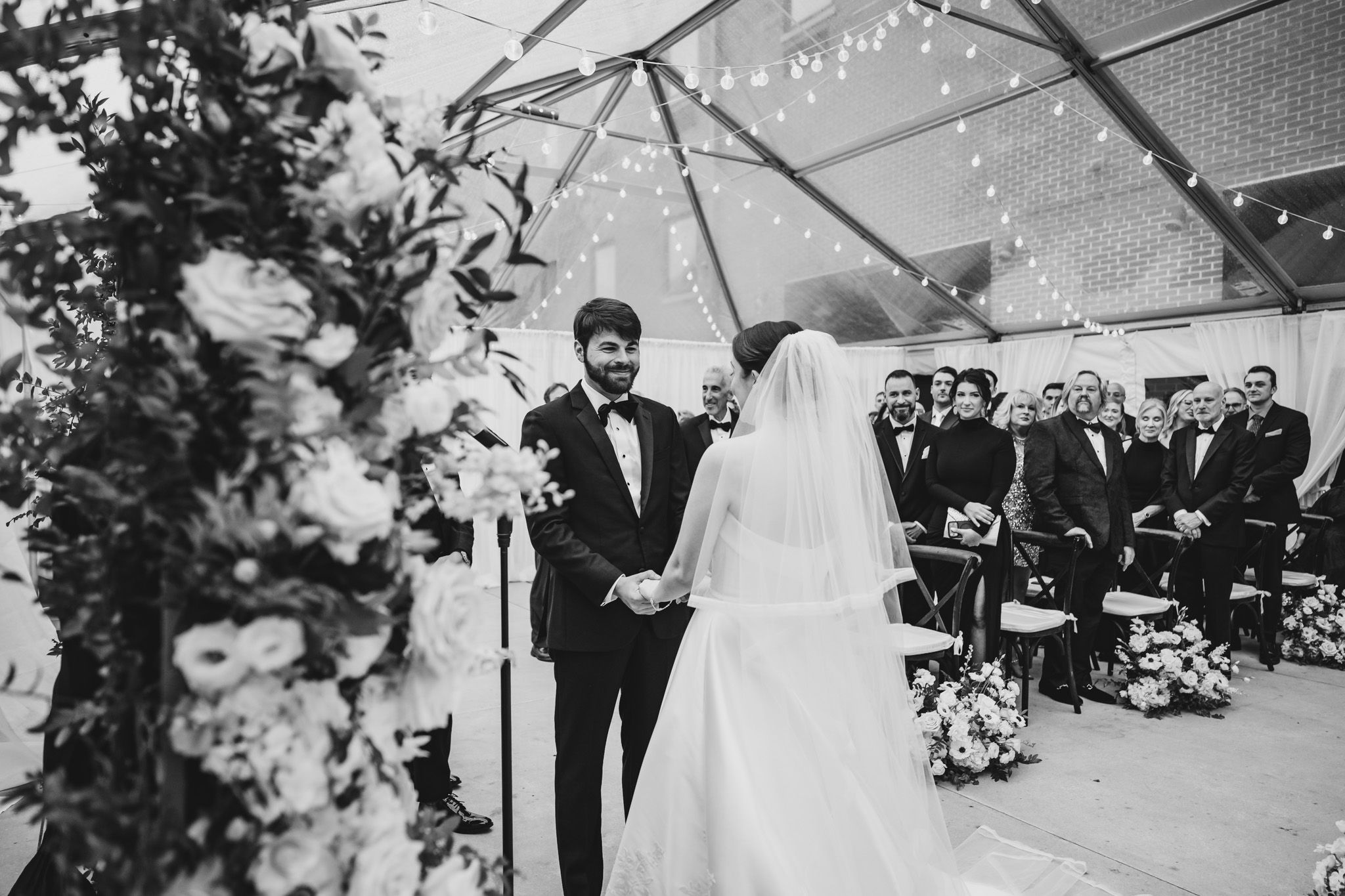 Black and white photo of groom smiling as bride walks down the aisle at floral-adorned altar.