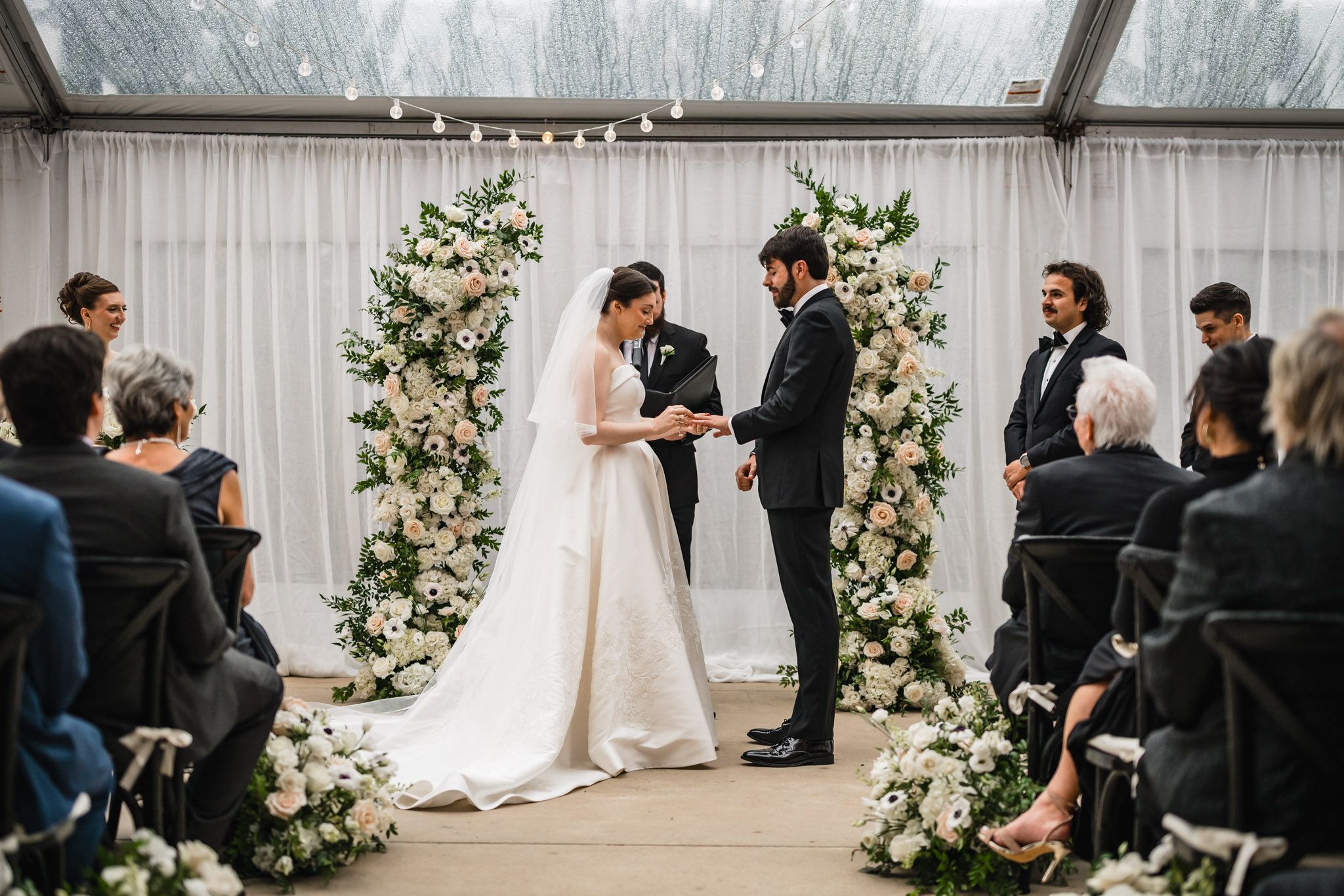 Bride and groom exchange vows at indoor ceremony flanked by tall white floral arrangements under a clear tent.