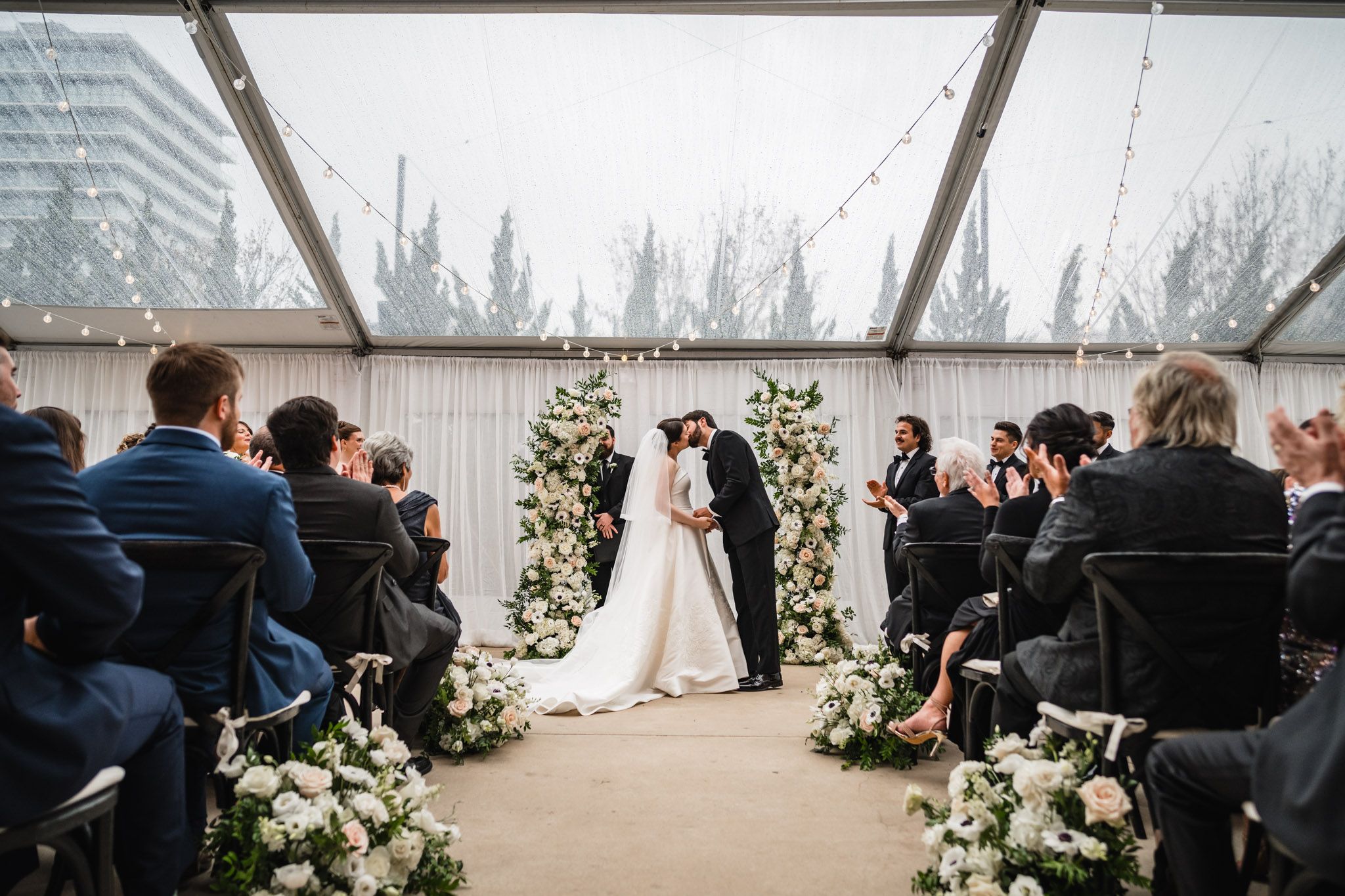 Couple sharing first kiss at altar surrounded by white floral arrangements under glass tent at wedding.