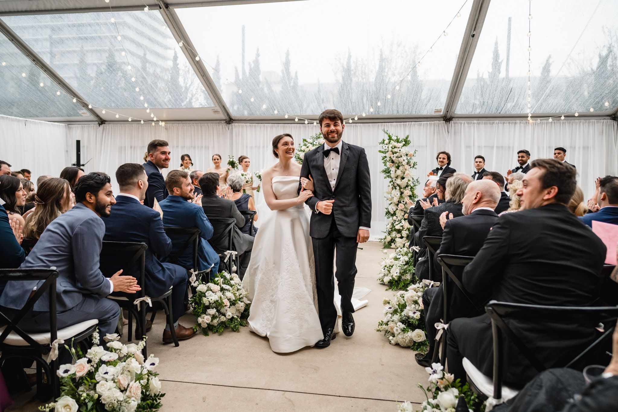 Bride and groom walk down the aisle under a clear tent adorned with string lights and white floral arrangements.