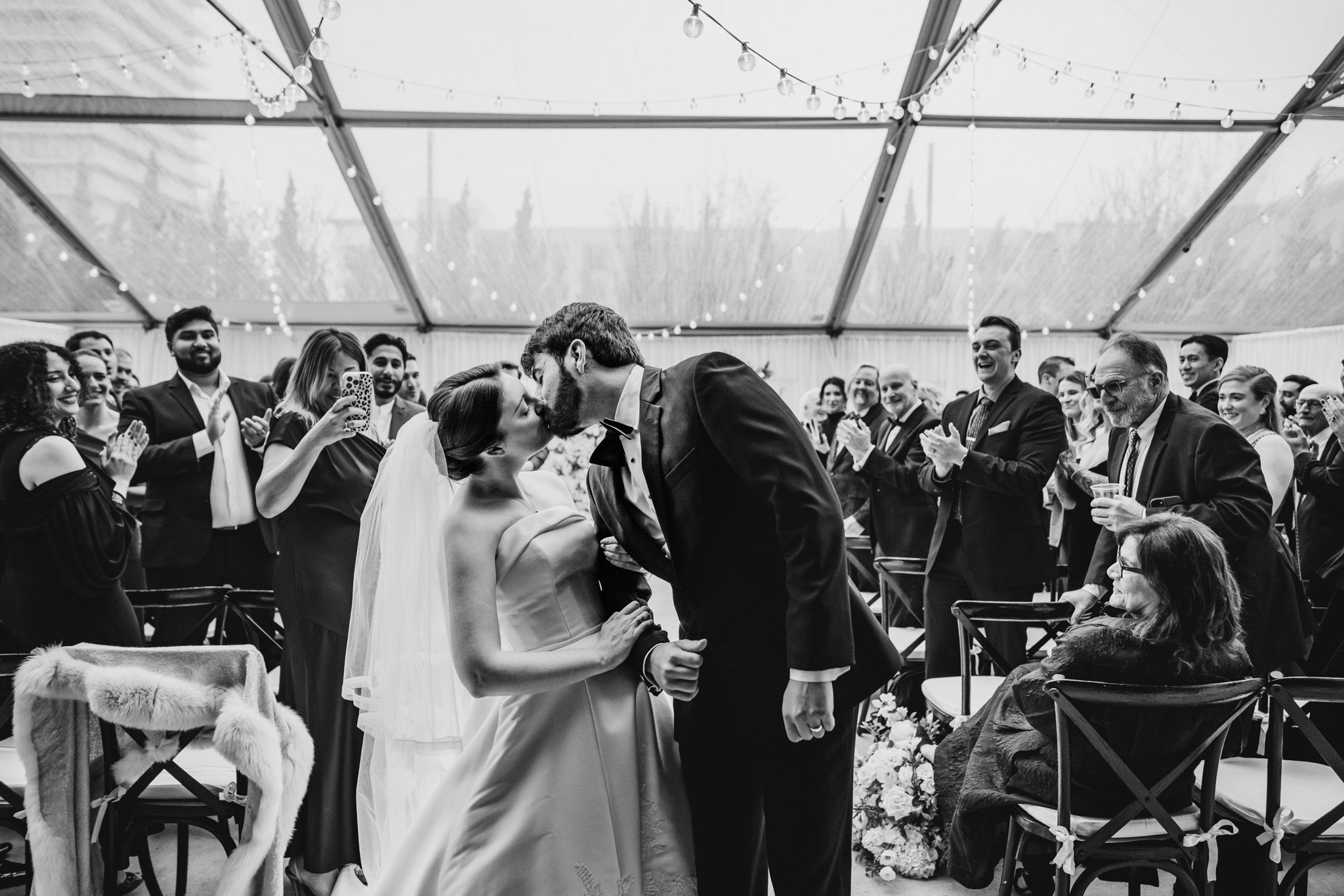 Bride and groom share their first kiss as newlyweds while guests cheer at their tented wedding reception.