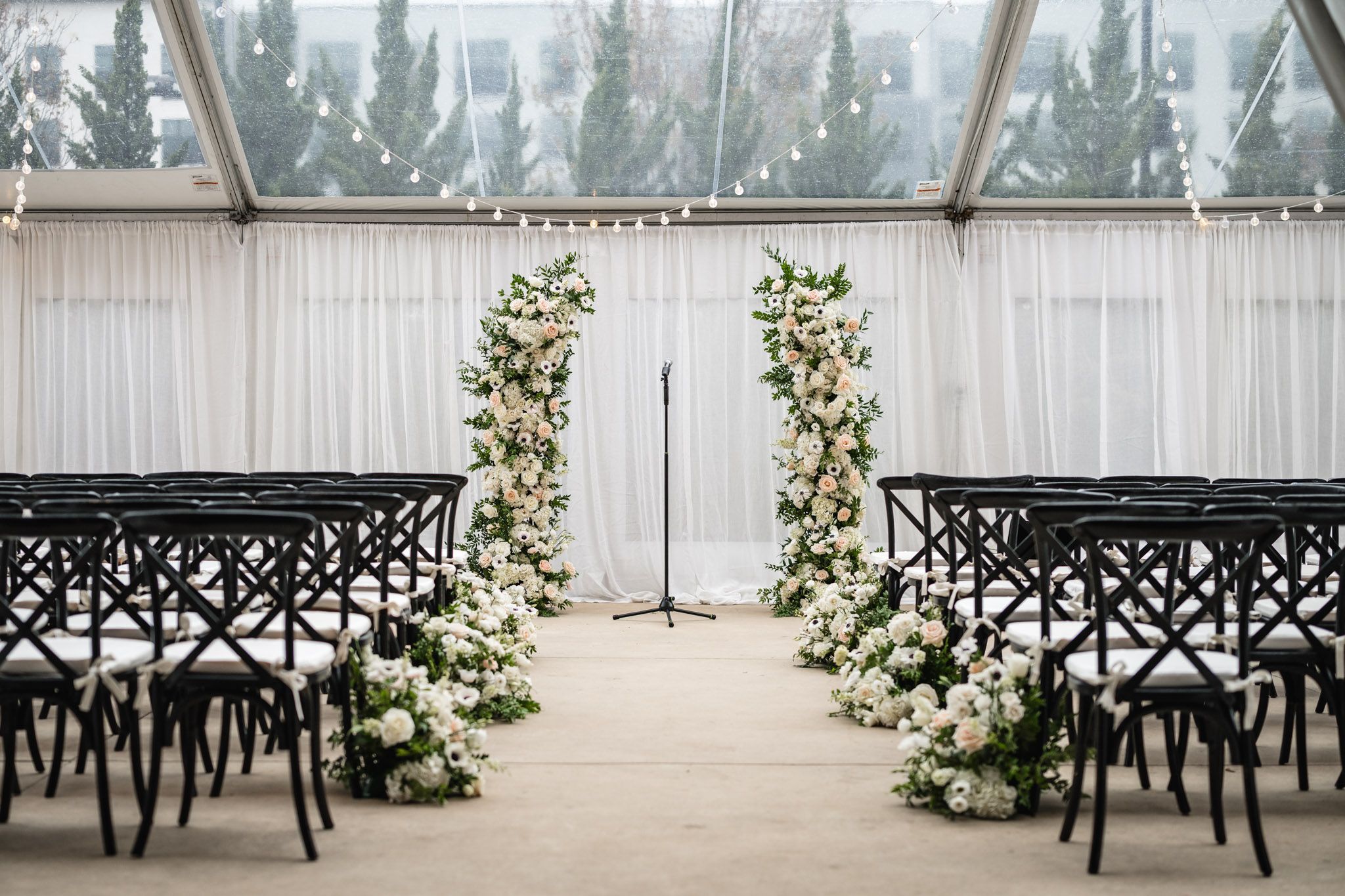 Wedding ceremony setup with tall white floral arch, black chairs, and string lights in glass tent.