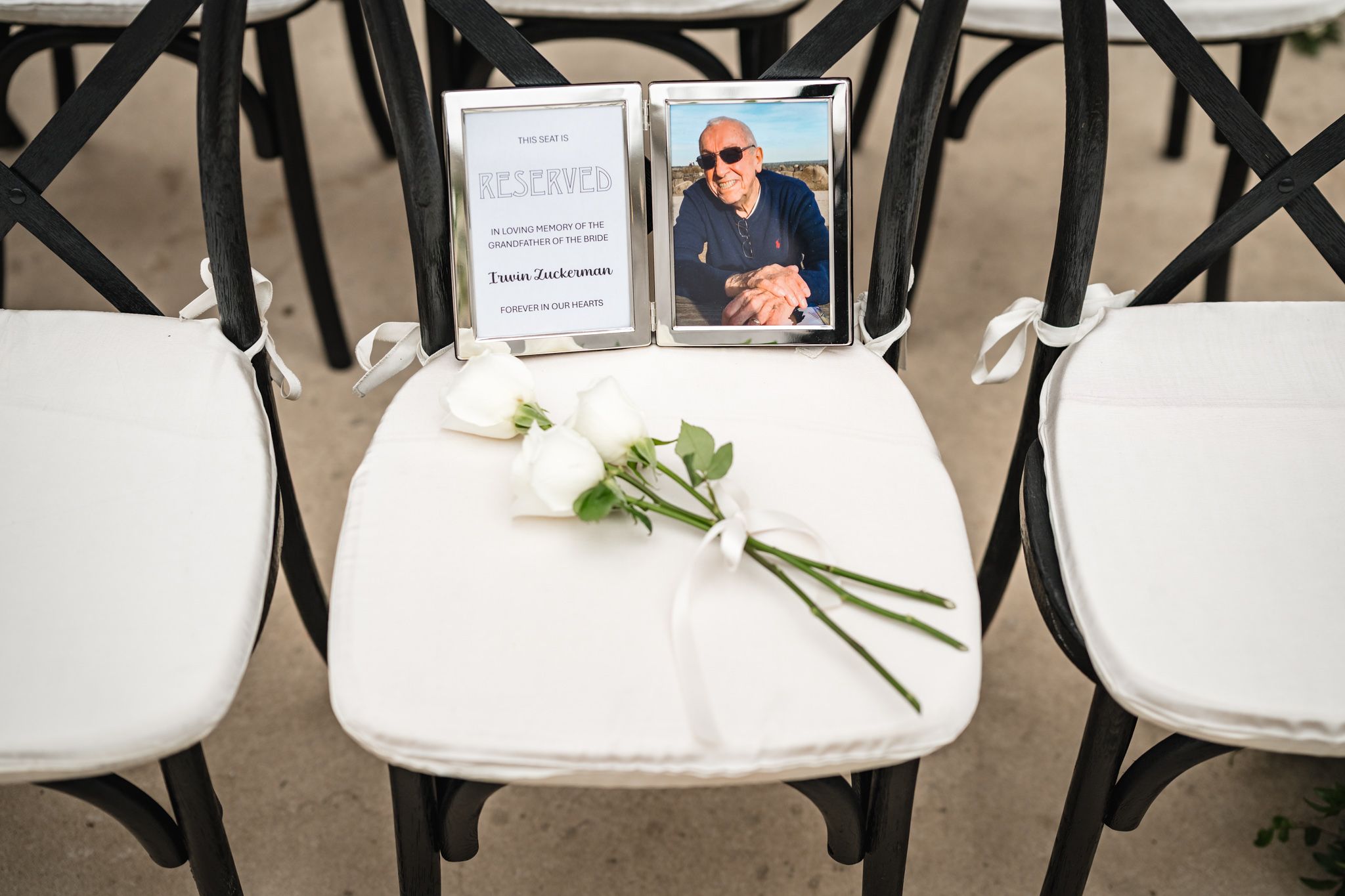 Memorial chair with white roses and framed photo honoring absent loved one at outdoor wedding ceremony.