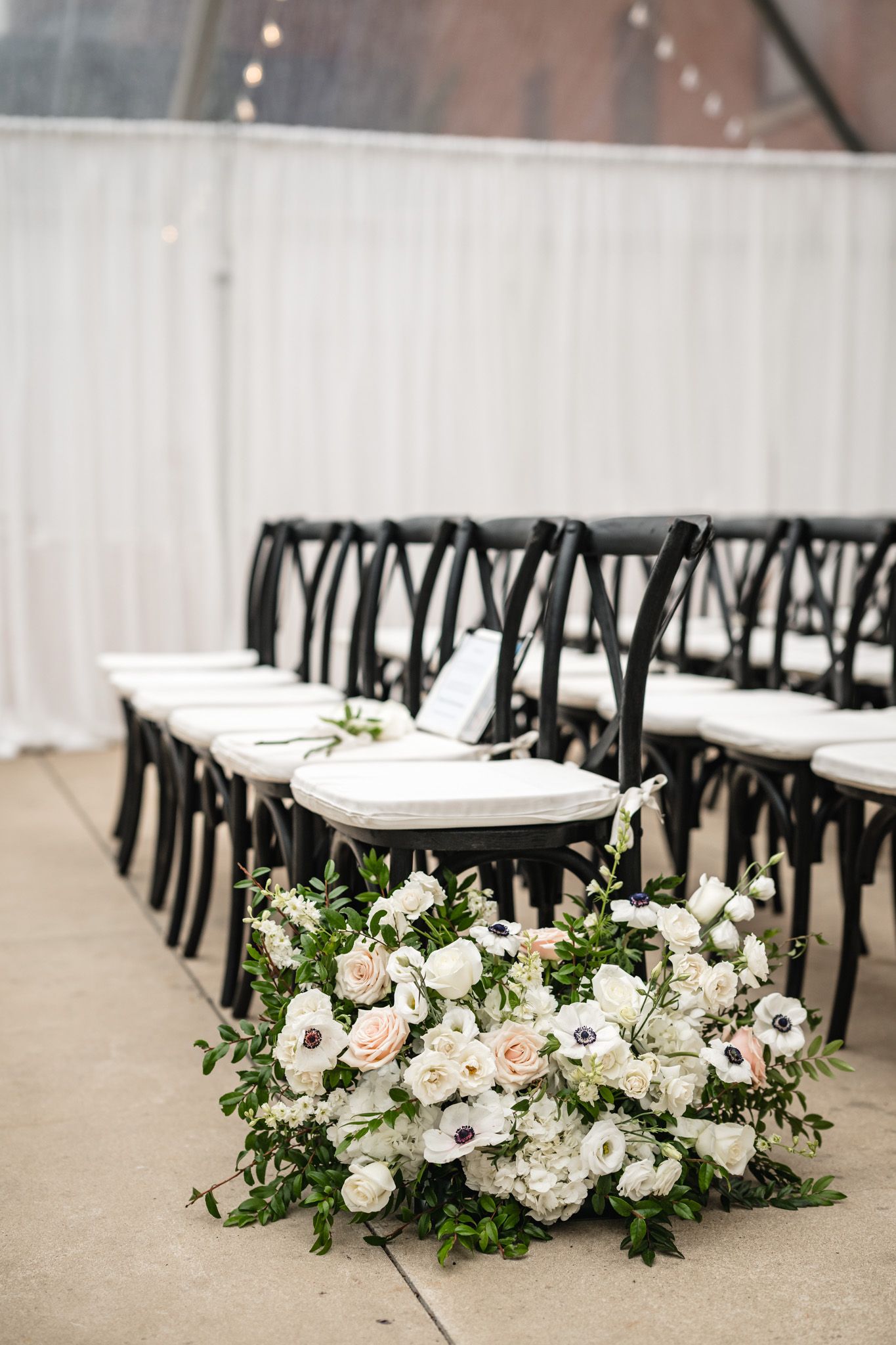 Black cross-back chairs with white cushions line a wedding ceremony aisle beside a lush white floral arrangement.