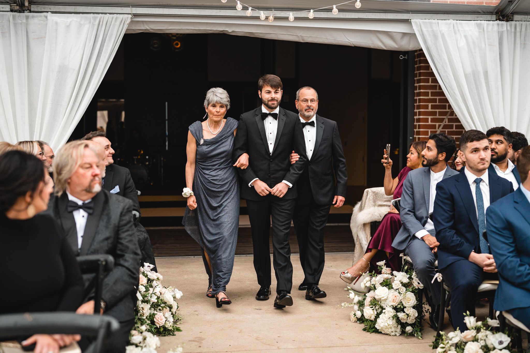 Groom in tuxedo walks down the aisle escorted by parents during an outdoor wedding ceremony.