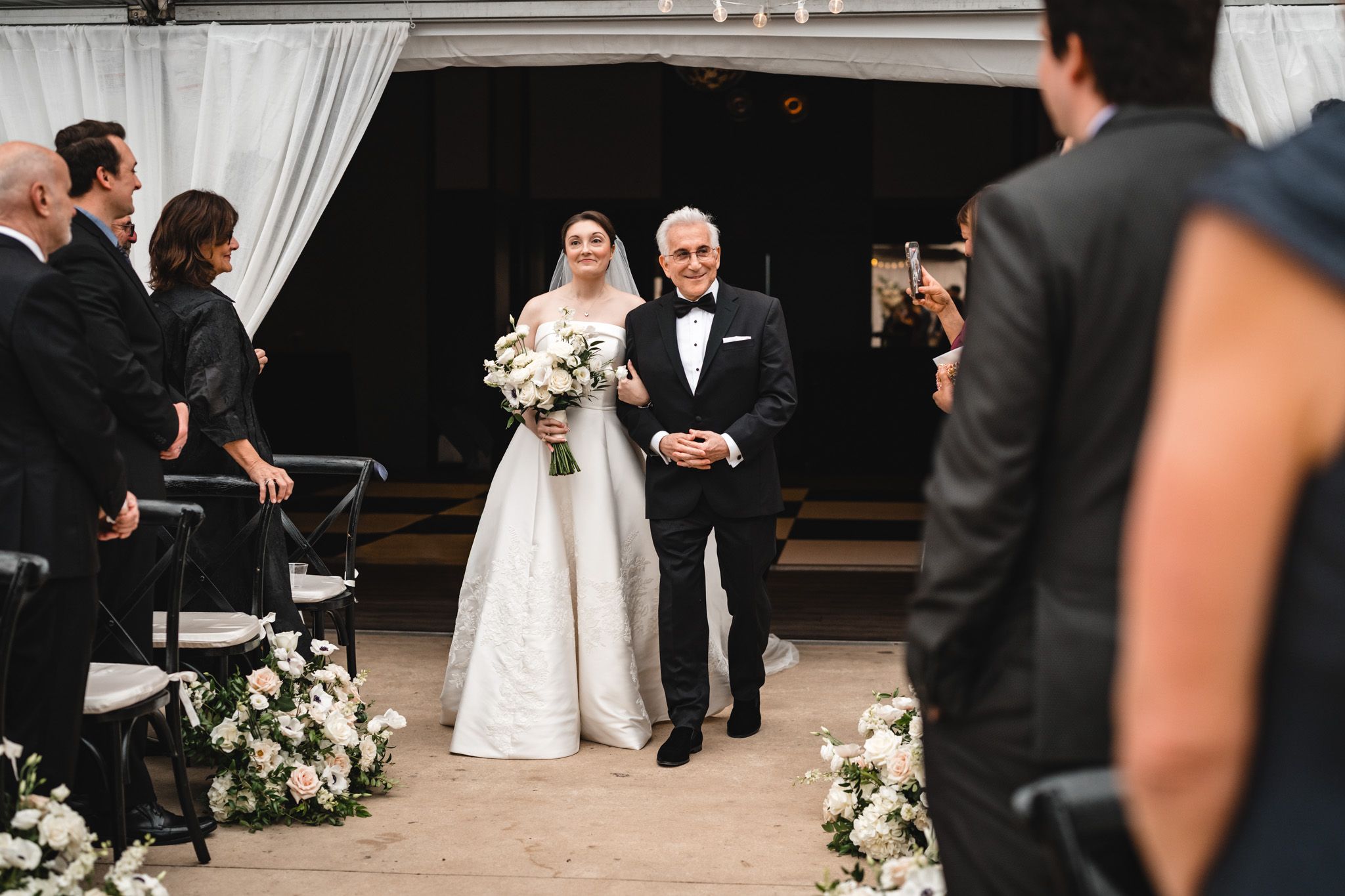 Bride walks down the aisle with her father at an elegant outdoor wedding ceremony with white floral arrangements.