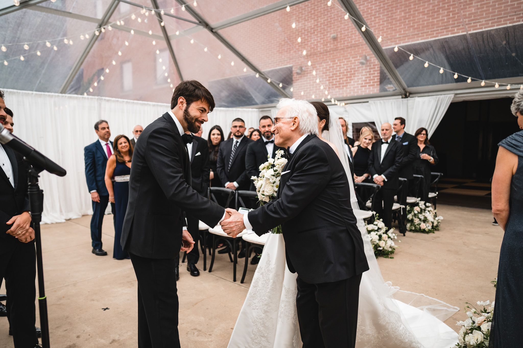 Groom shaking hands with father of bride during wedding ceremony under elegant clear tent with string lights.