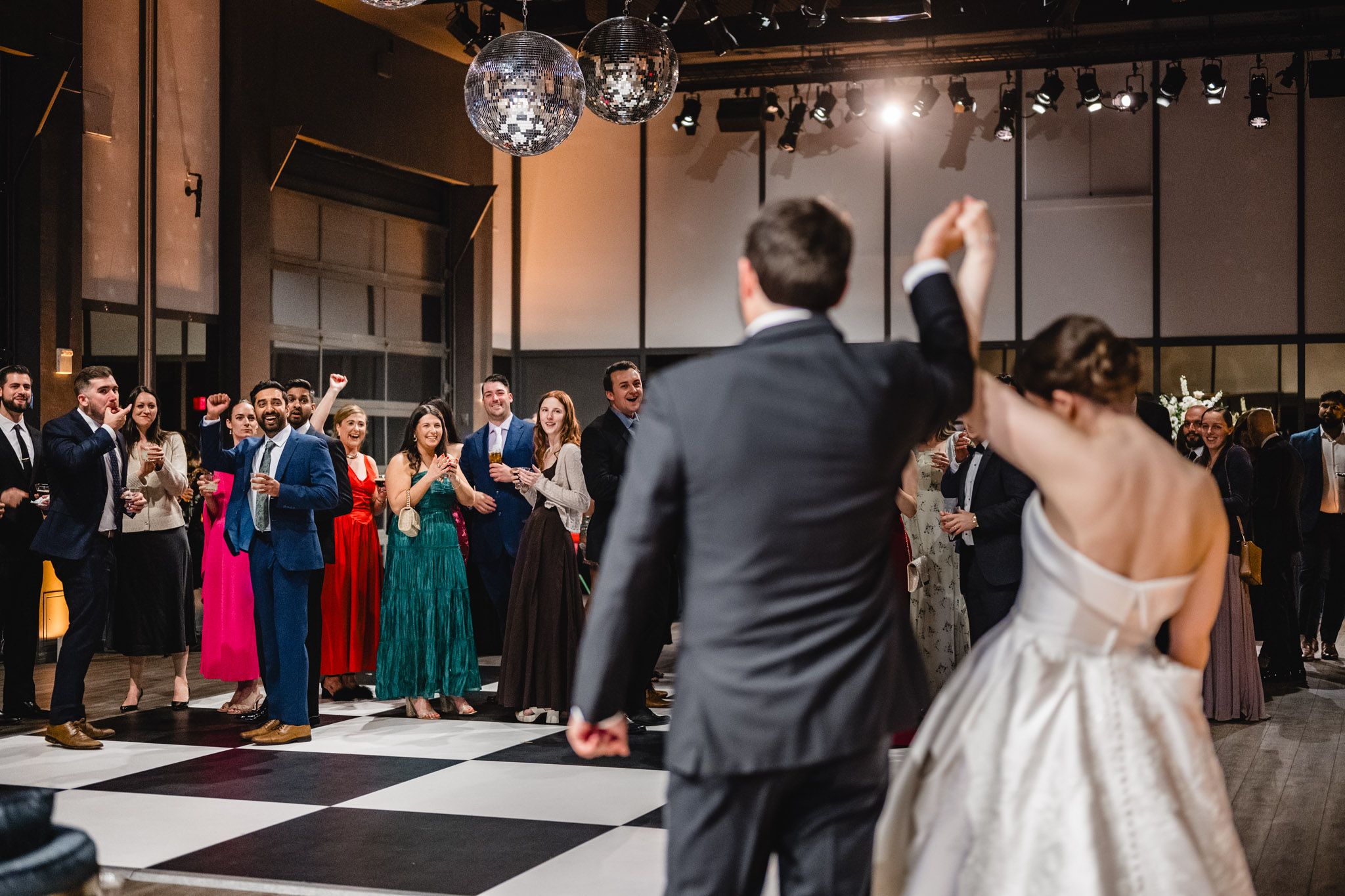 Bride and groom celebrate their first dance on a black and white checkered floor surrounded by cheering guests.