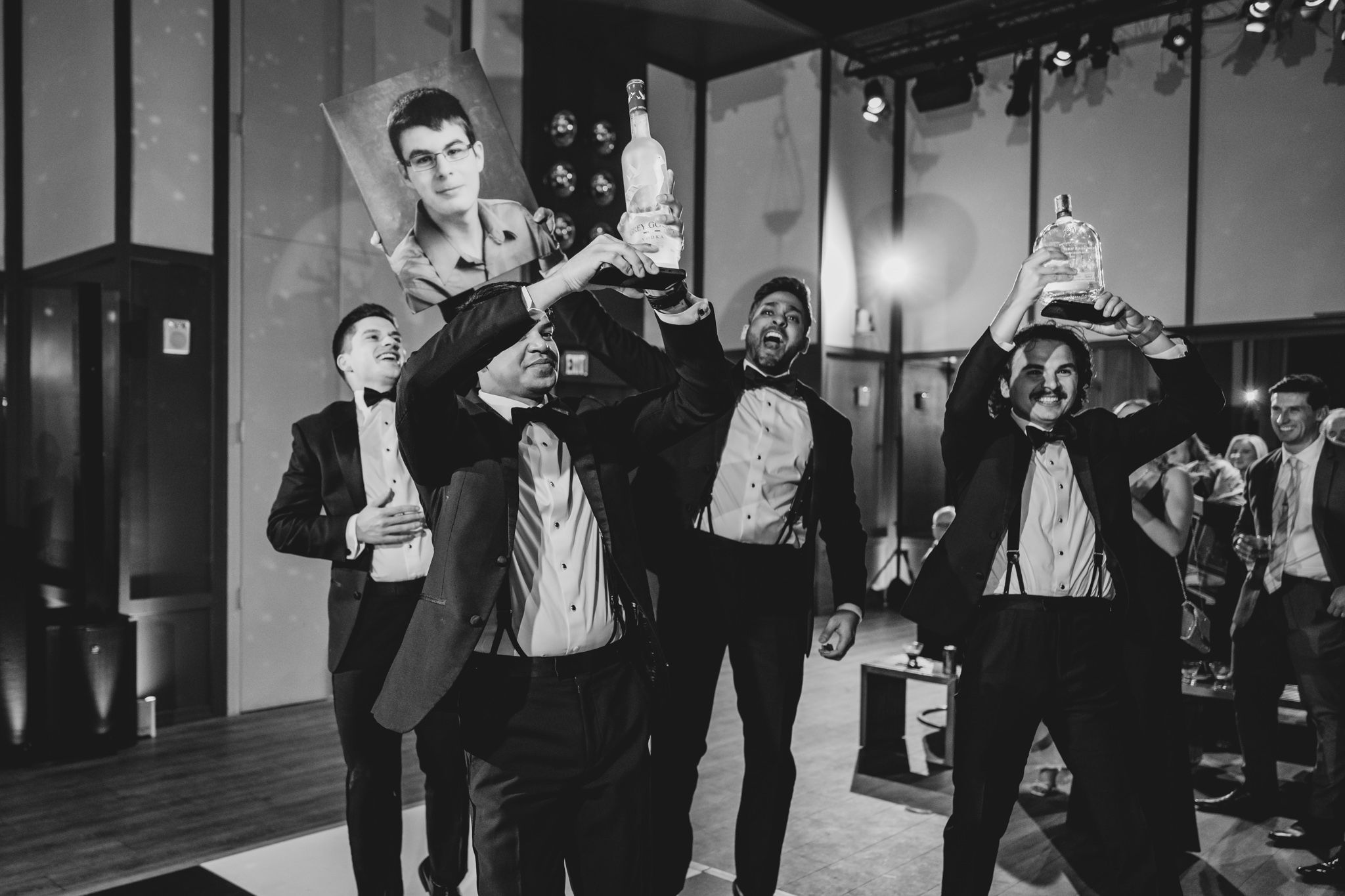 Groomsmen celebrate at a wedding reception, holding bottles and lifting the groom on their shoulders.