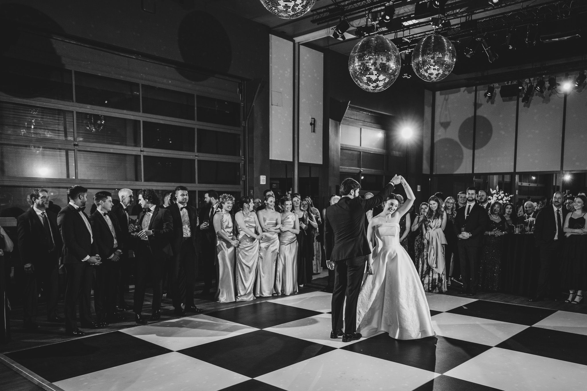 Black and white photo of bride and groom first dance on checkered floor with guests watching.
