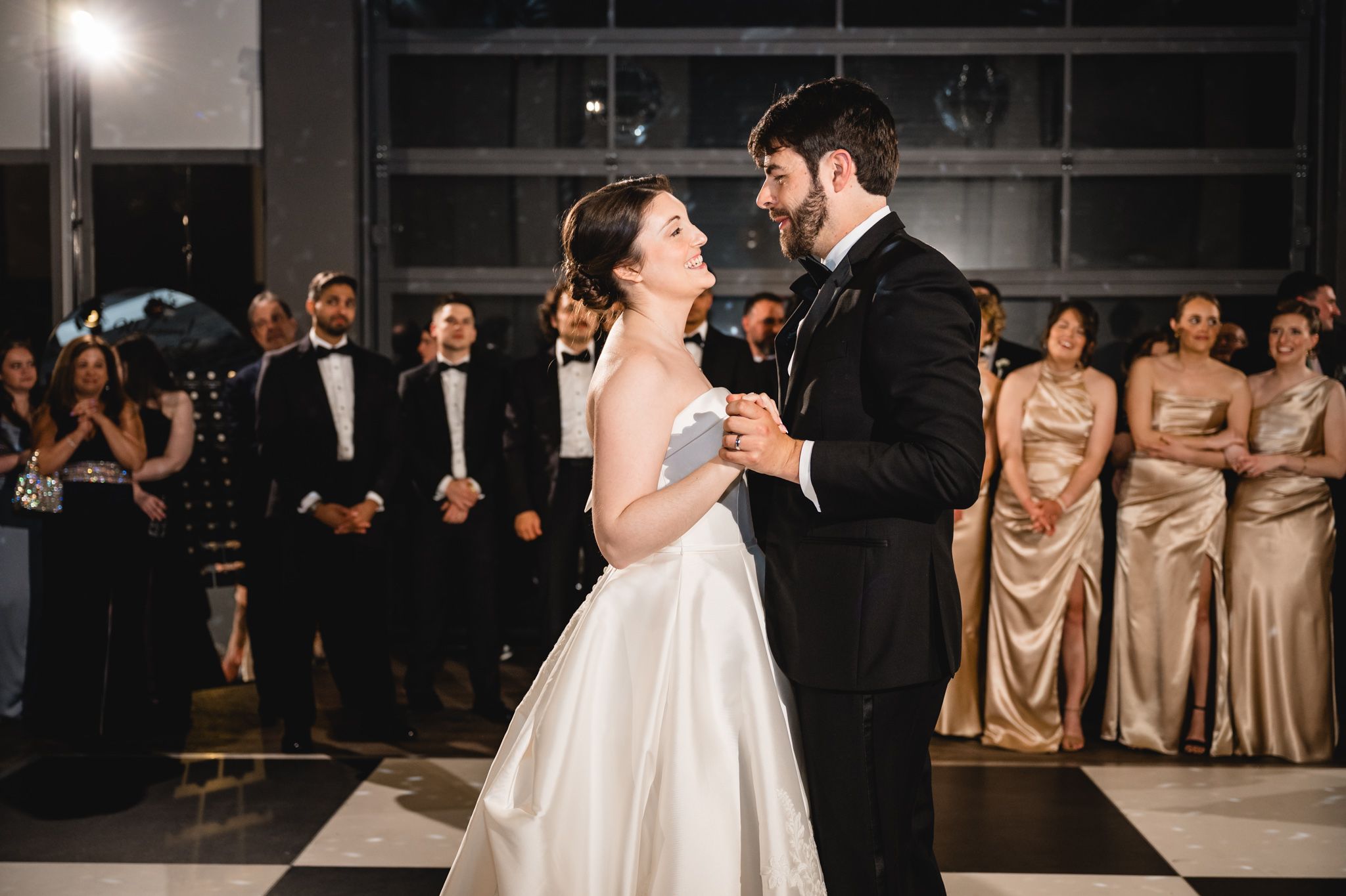 Bride and groom share their first dance on a black and white checkered floor surrounded by wedding guests.