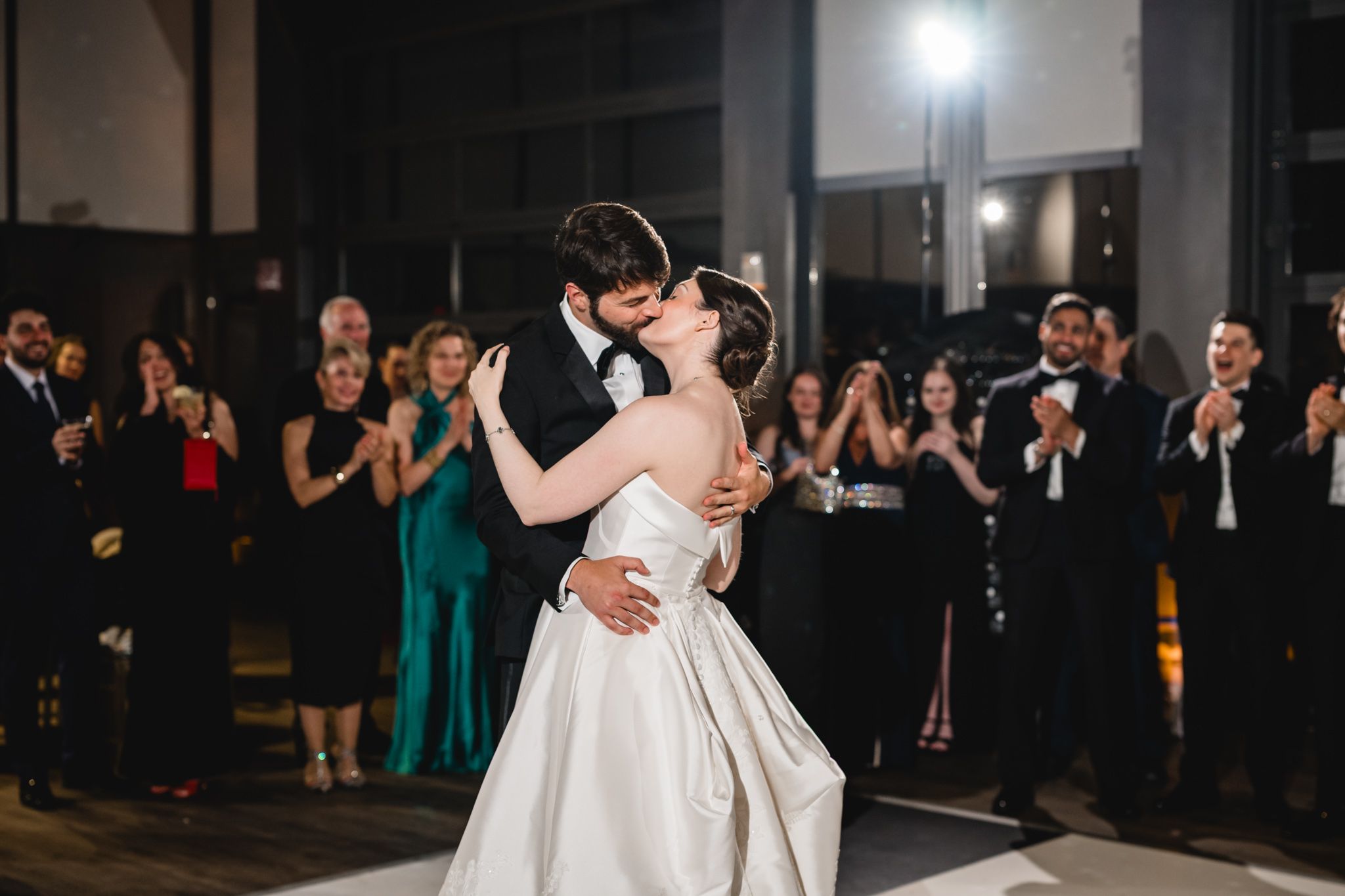 Bride and groom share passionate first dance kiss while guests applaud in dimly lit reception venue.