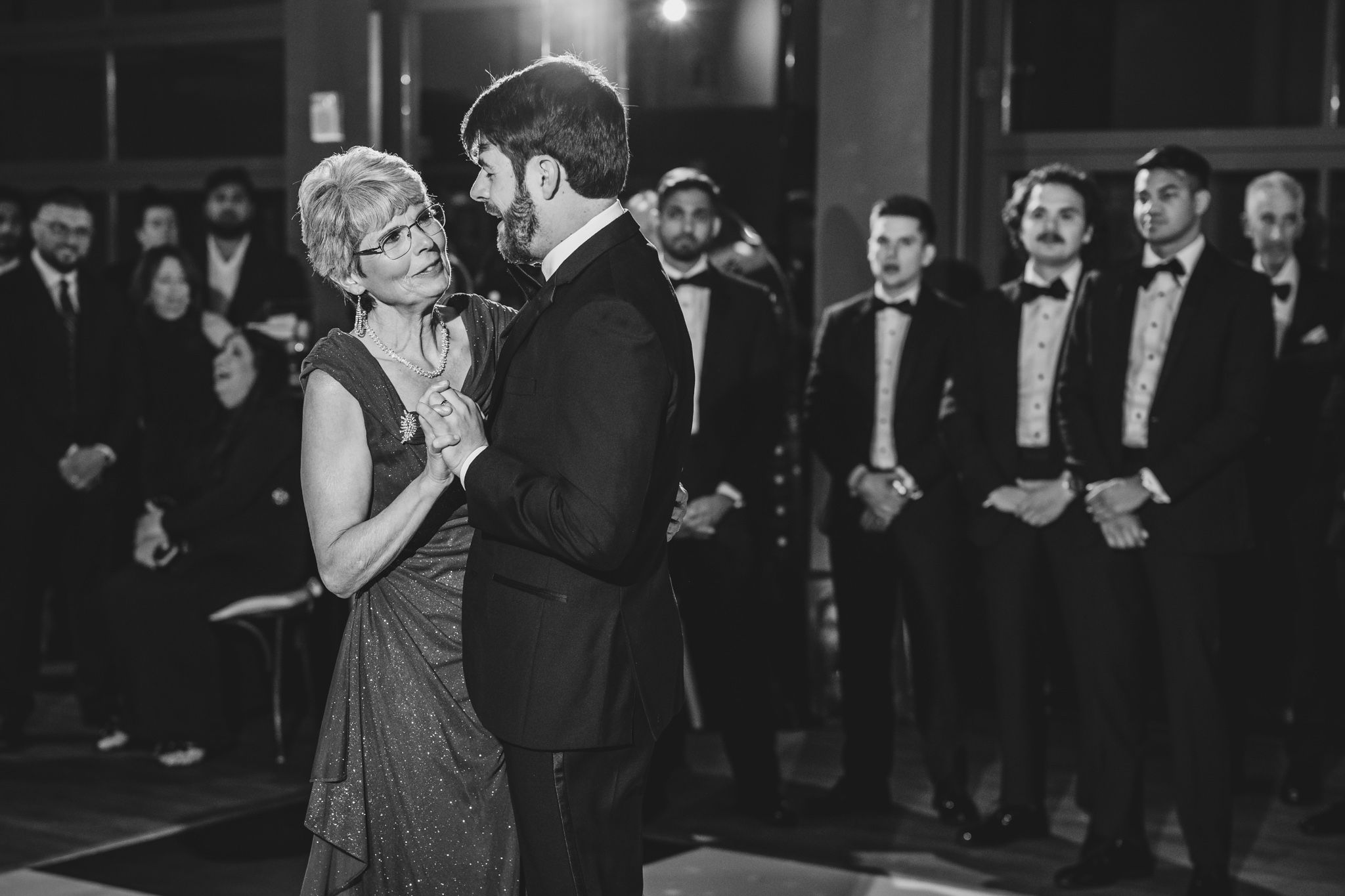 Groom shares emotional mother-son dance in black and white photo as guests watch at wedding reception.