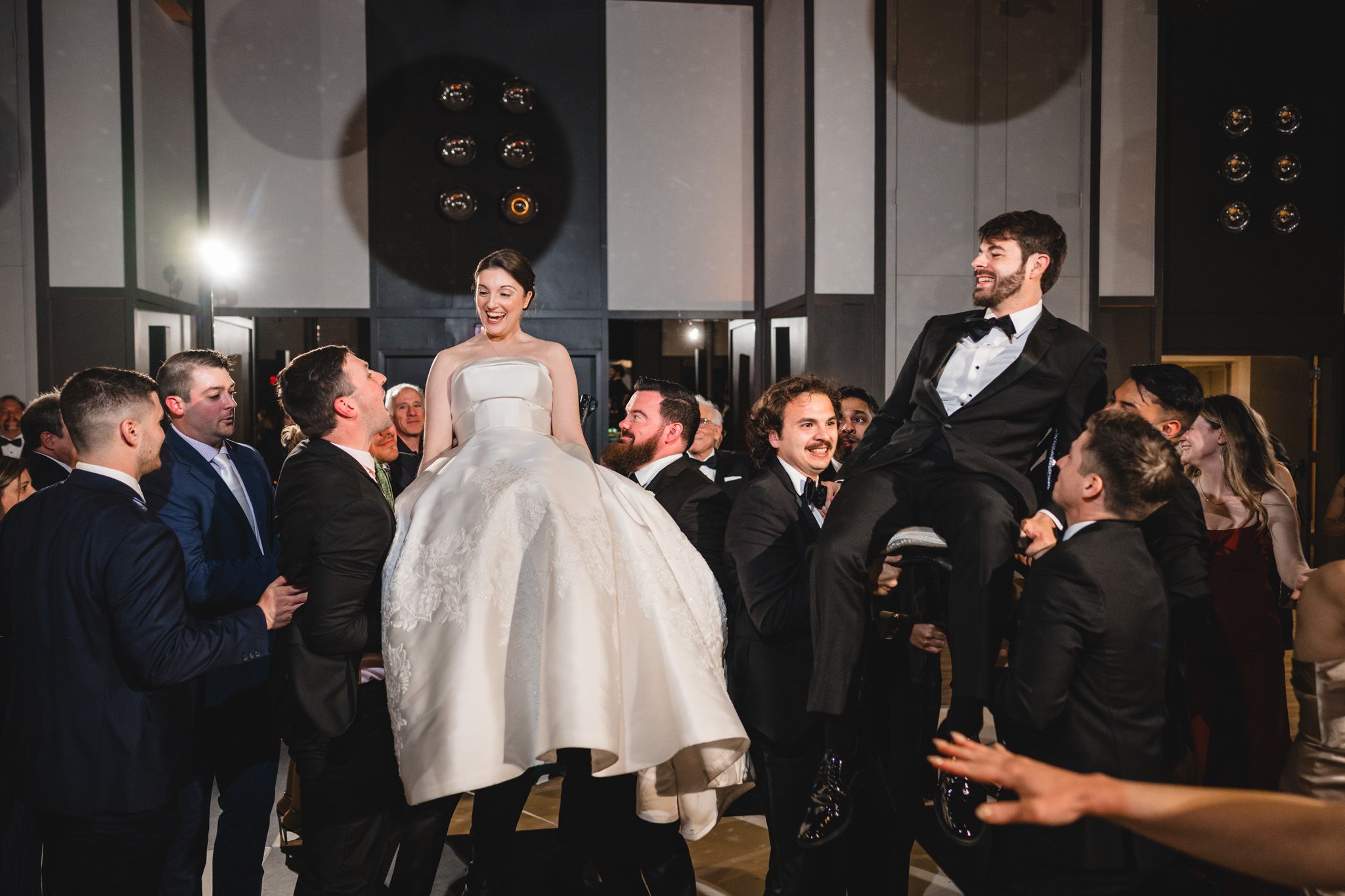 Bride and groom lifted by guests during joyful Jewish wedding hora dance celebration.