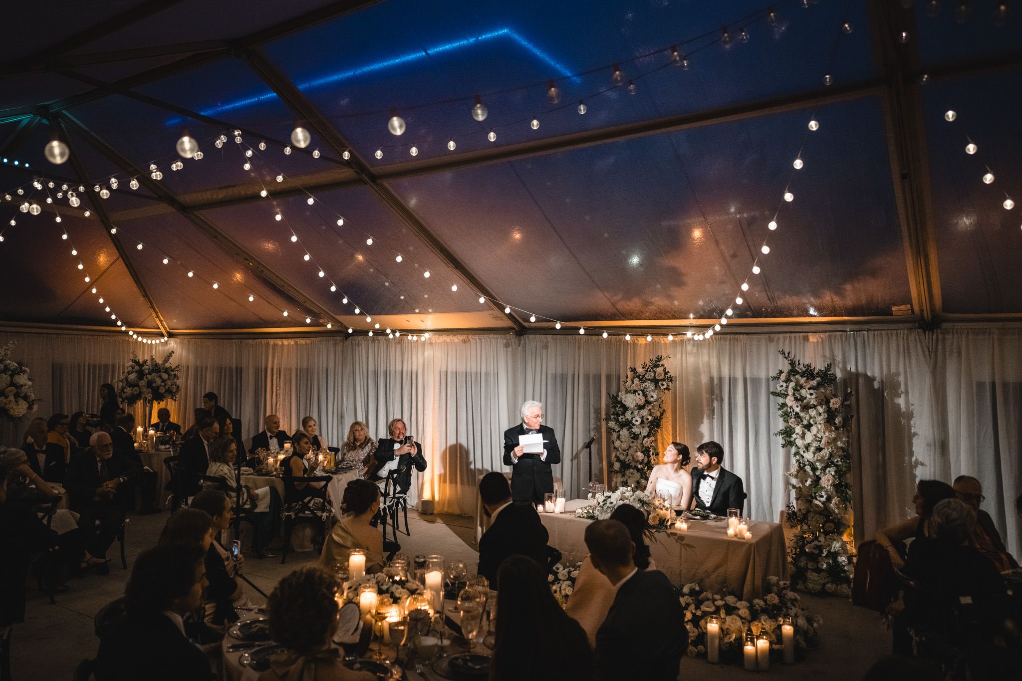 Wedding guests gather under string lights as someone delivers a toast at the candlelit sweetheart table.