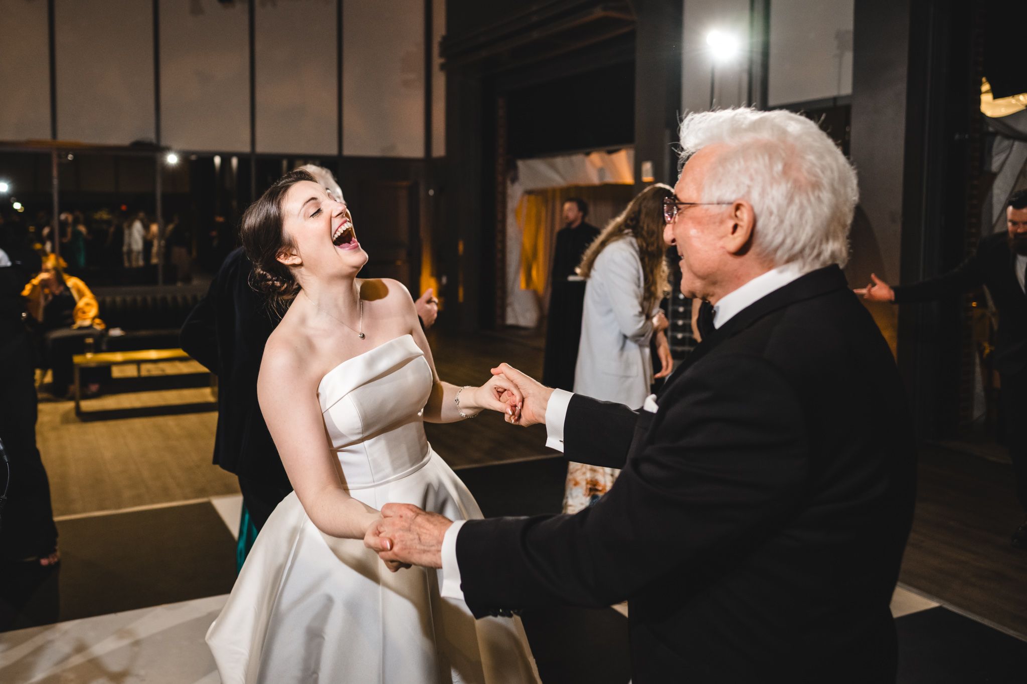 Bride laughs joyfully while dancing with an older gentleman in a tuxedo at her wedding reception.