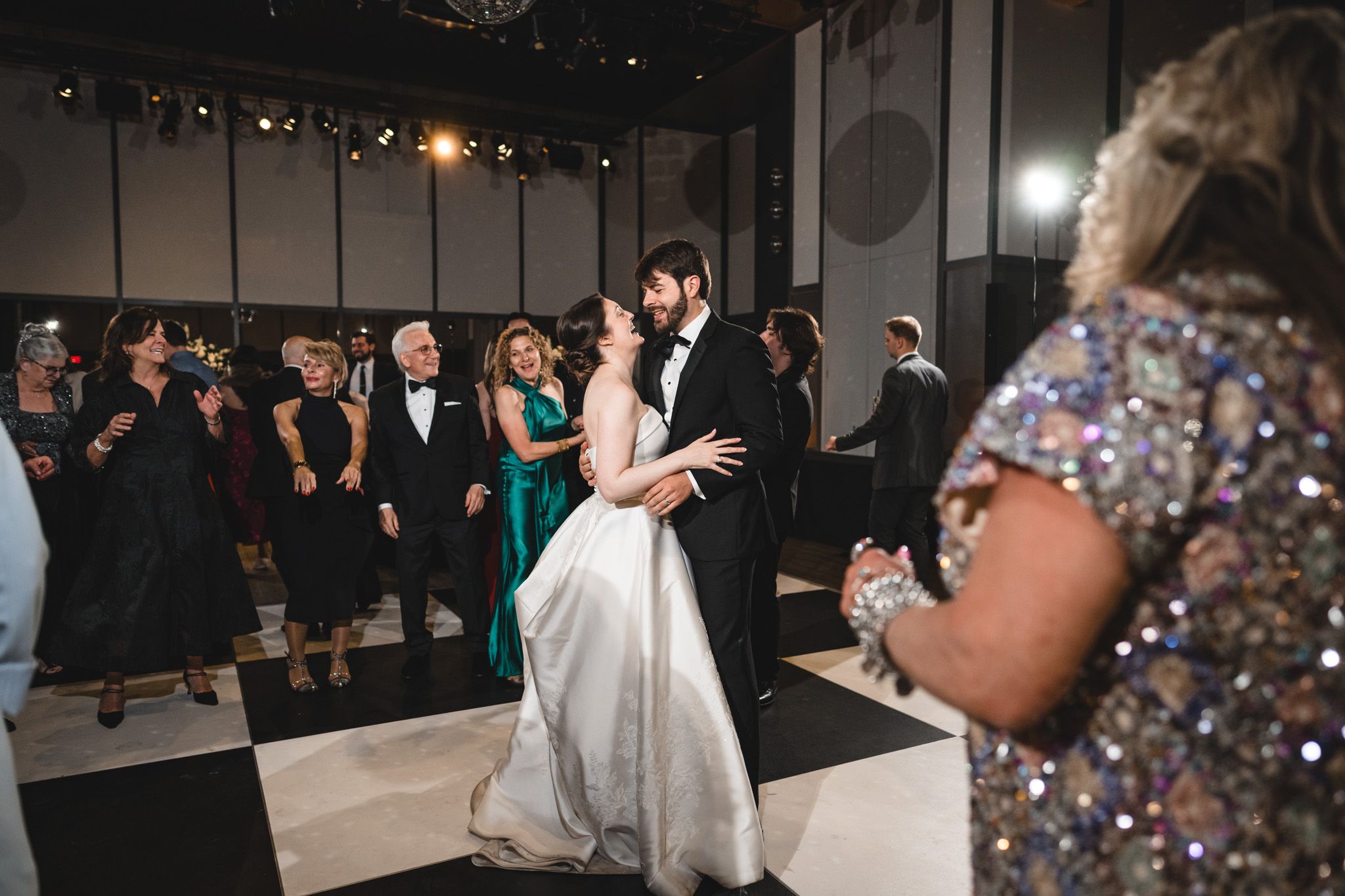 Newlyweds share a romantic first dance on a black and white floor surrounded by smiling wedding guests.