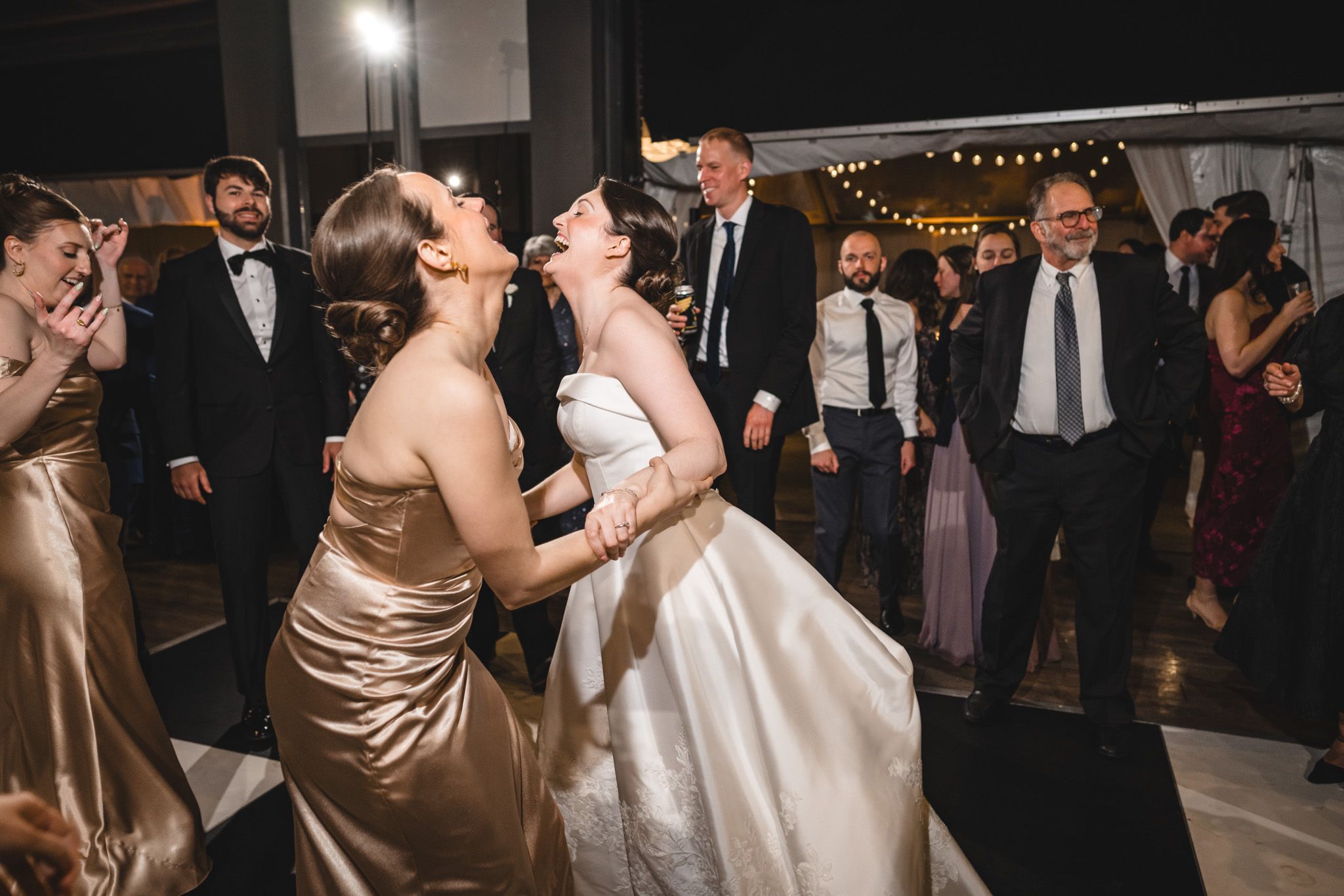 Joyful bride dancing with bridesmaid in gold dress surrounded by wedding guests on the reception dance floor.