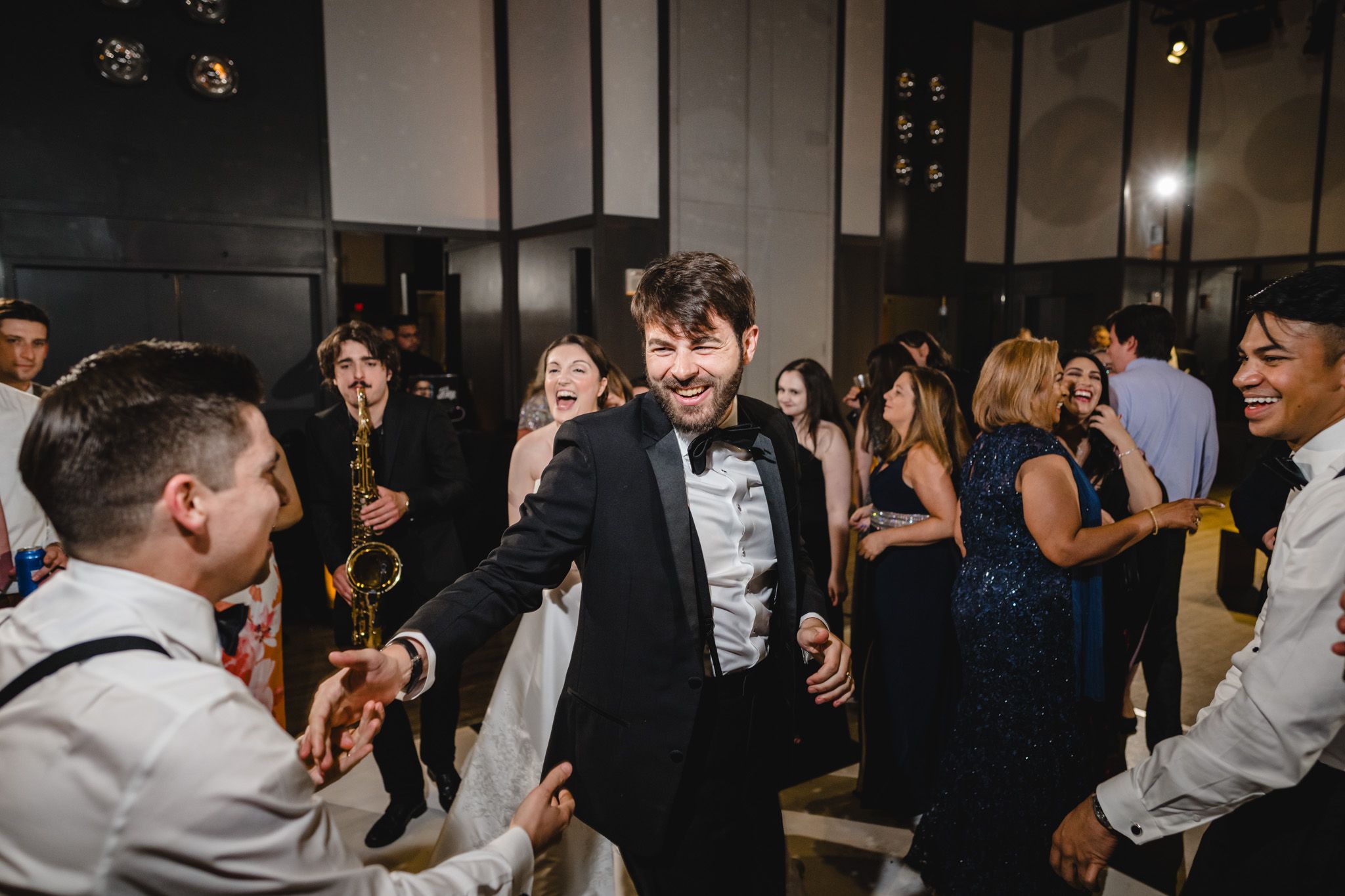Groom celebrates joyfully on the dance floor with guests and live saxophonist at wedding reception.