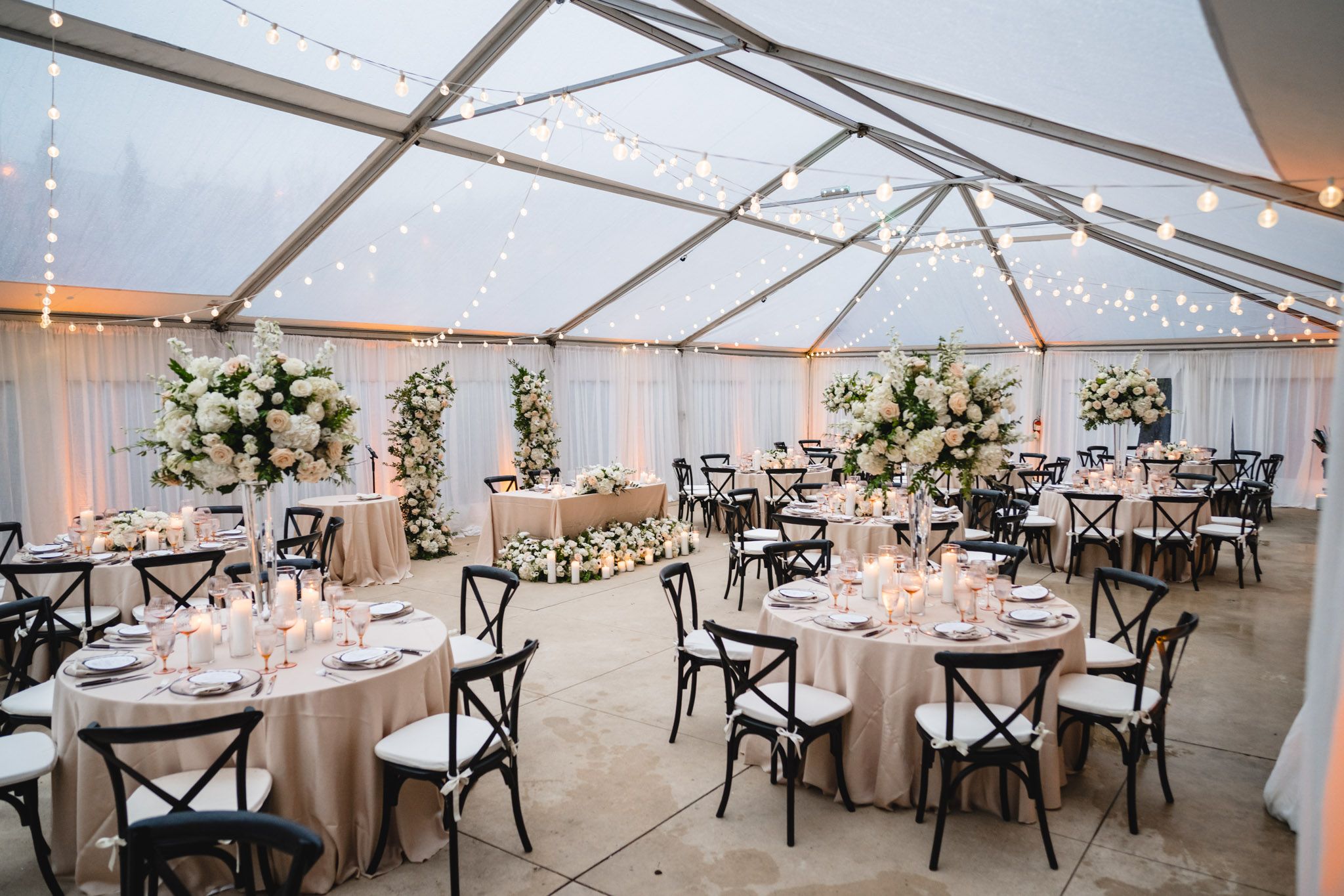 Elegant tented wedding reception with round tables, string lights, cross-back chairs, and white floral centerpieces.