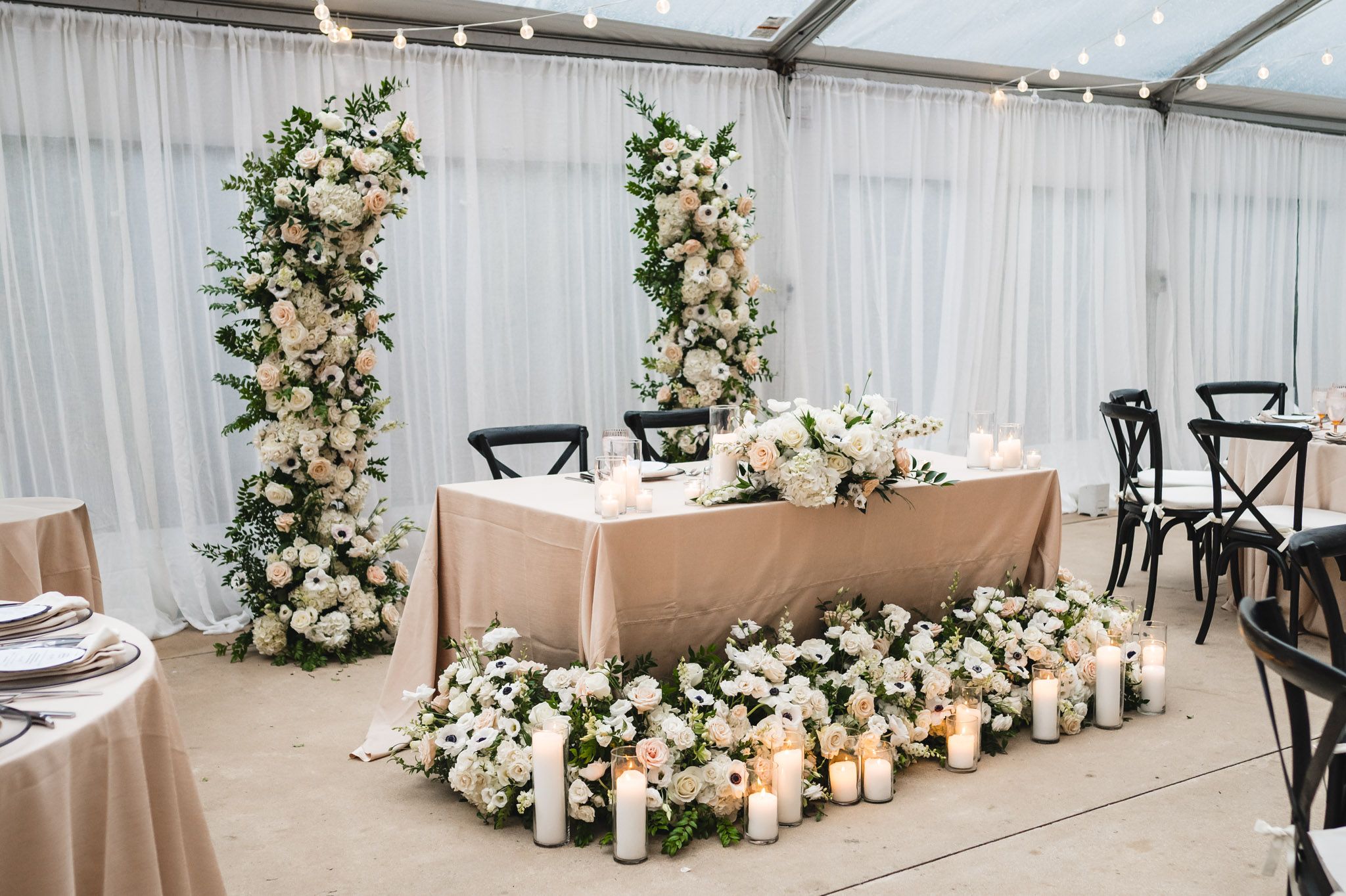 Sweetheart table adorned with lush white floral columns, candles, and garden flowers at a tented wedding reception.