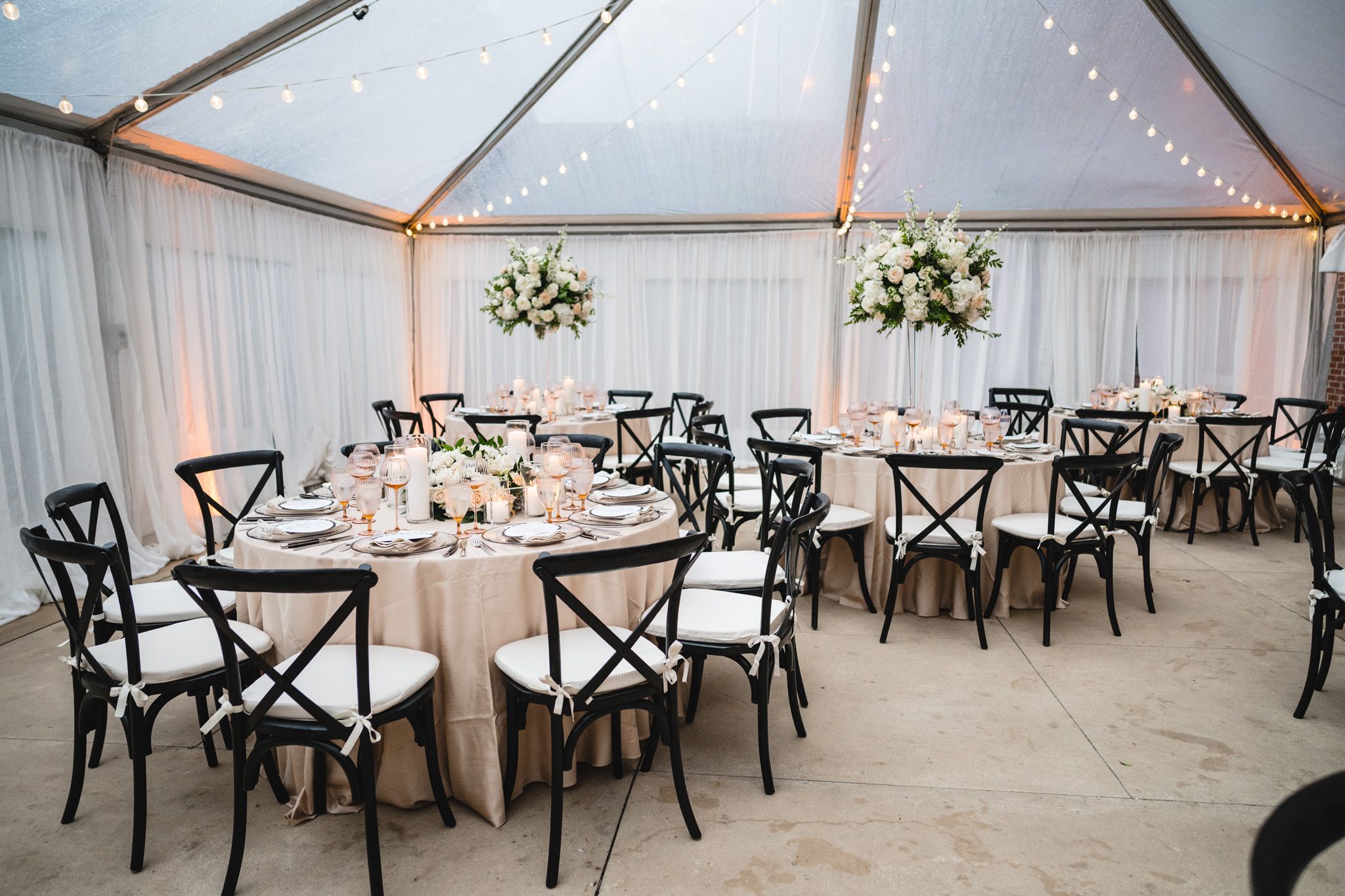 Elegant wedding reception under a clear tent with black cross-back chairs, round tables, and tall white floral centerpieces.