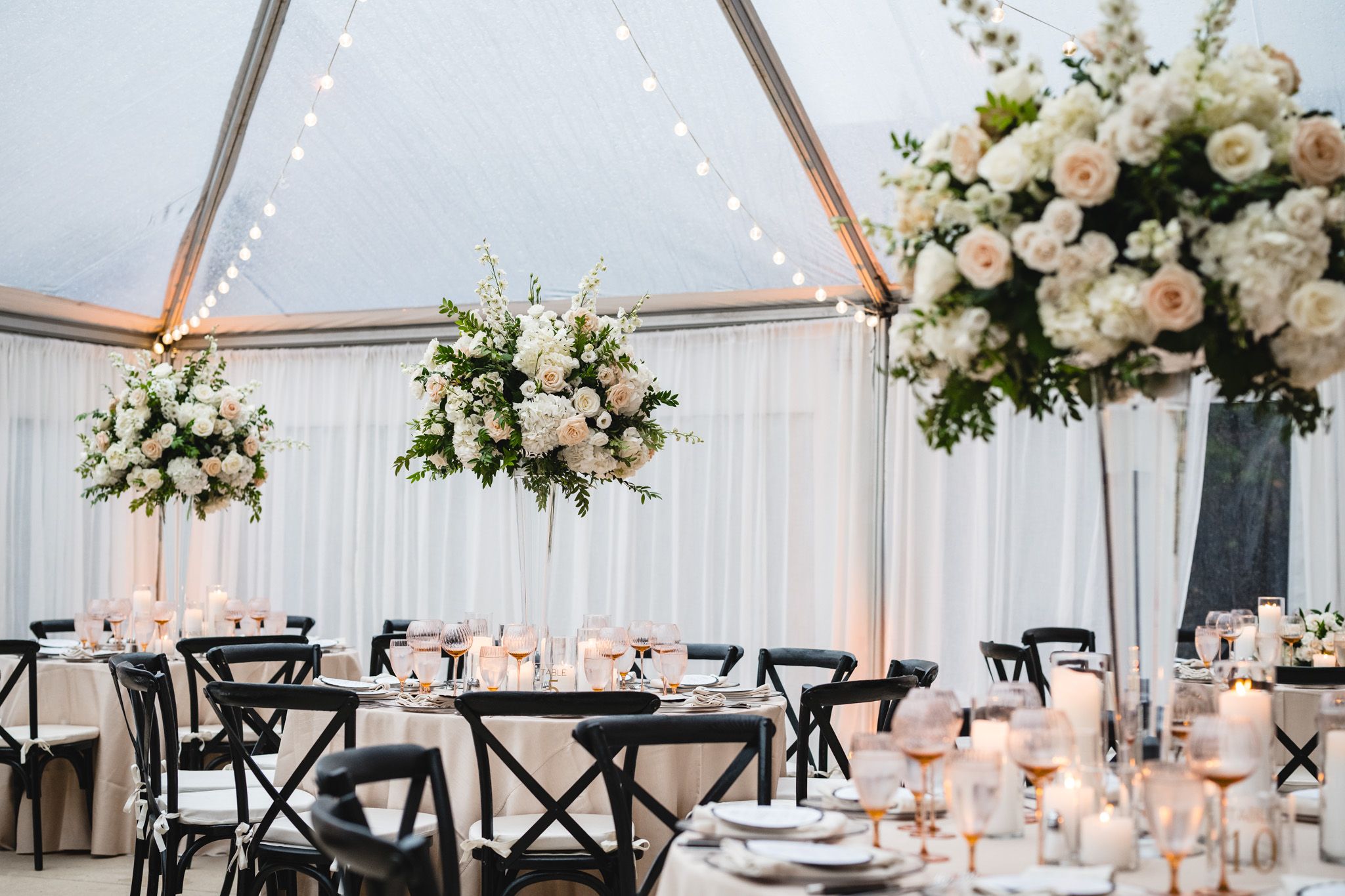 Elegant wedding reception under clear tent with tall floral centerpieces of white roses and candlelit tables.