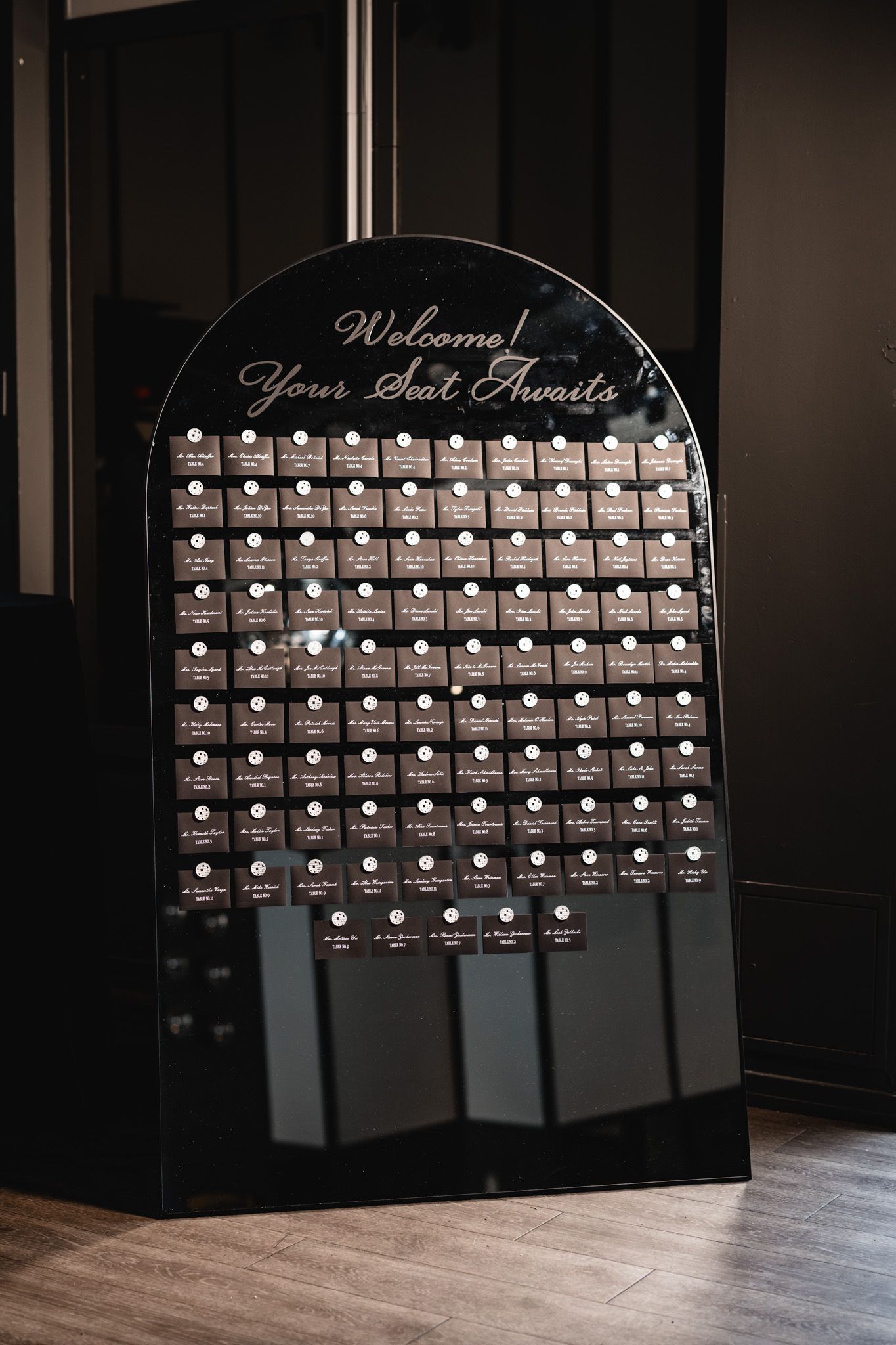 Black arched seating chart display with gold script reading Welcome Your Seat Awaits at a wedding venue.