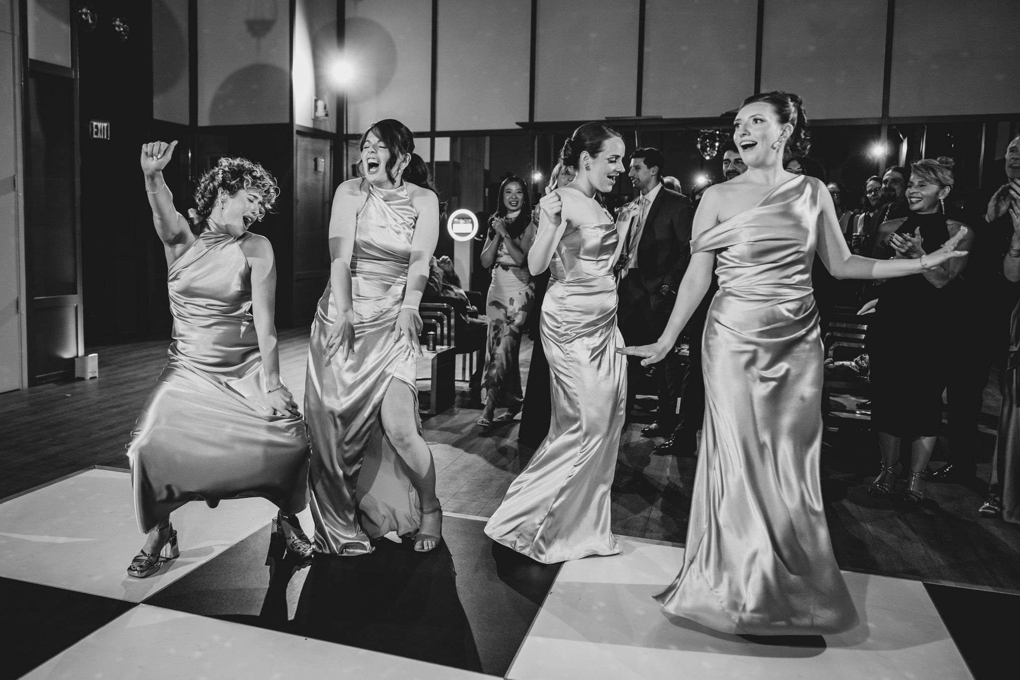 Four bridesmaids in silver satin gowns dance joyfully on a black and white checkered dance floor.
