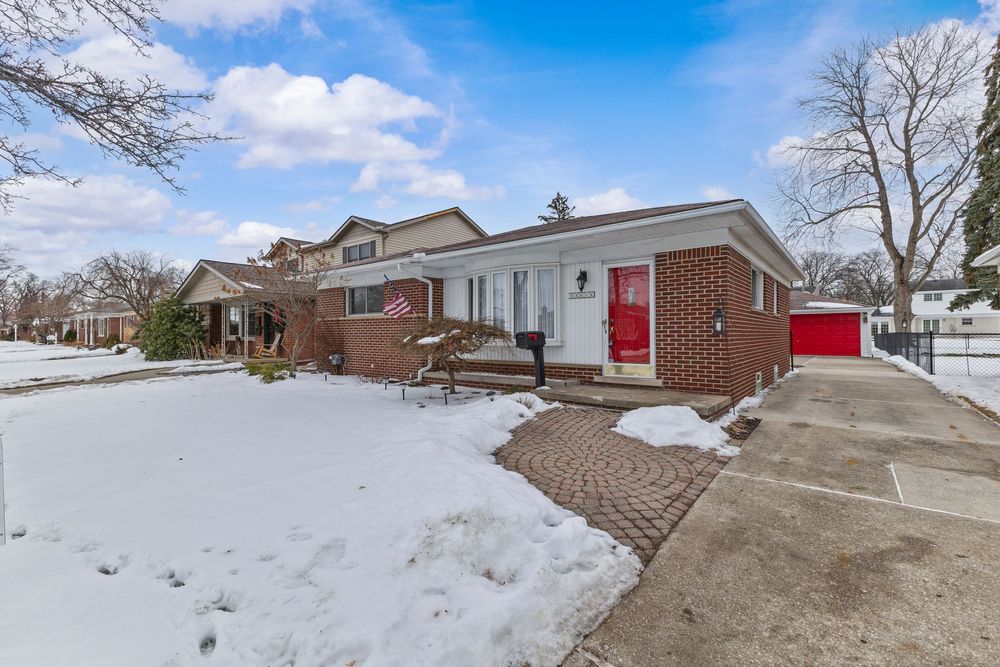 Brick ranch home with red door and attached garage, covered in snow during winter, featuring a brick paved walkway.