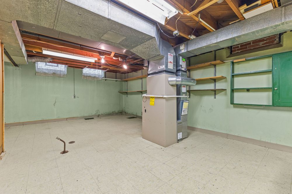 Unfinished basement with green painted walls, exposed ceiling, utility furnace, shelving units, and vinyl tile flooring.