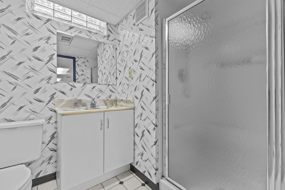 Bathroom with patterned wallpaper, white vanity with sink, and a glass-door walk-in shower on white tile flooring.
