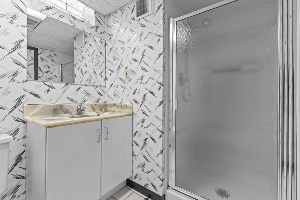 Bathroom with marble-patterned wallpaper, white vanity with sink, large mirror, and glass-enclosed shower stall.