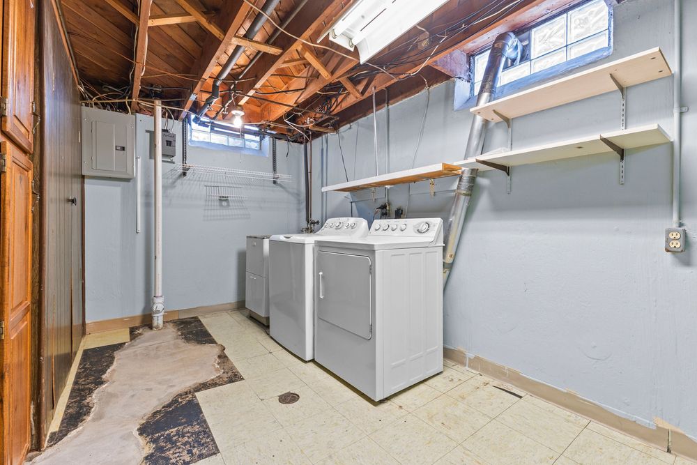 Unfinished basement laundry room with washer and dryer, exposed wooden ceiling beams, shelving, and tiled flooring.
