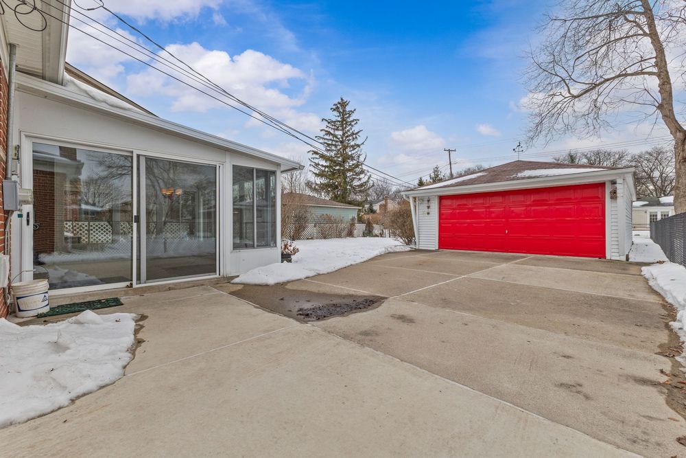 Suburban home exterior with sunroom, red garage door, and concrete driveway dusted with snow on a clear winter day.