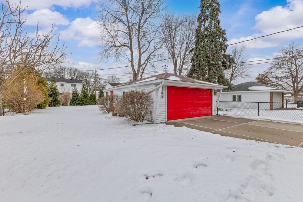 Snow-covered yard with detached garage featuring bold red doors and evergreen trees in winter.