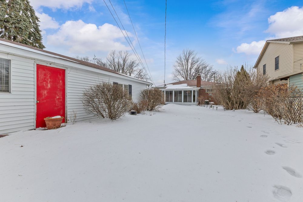 Snow-covered backyard with red side door, bare shrubs, and sunroom addition in winter.