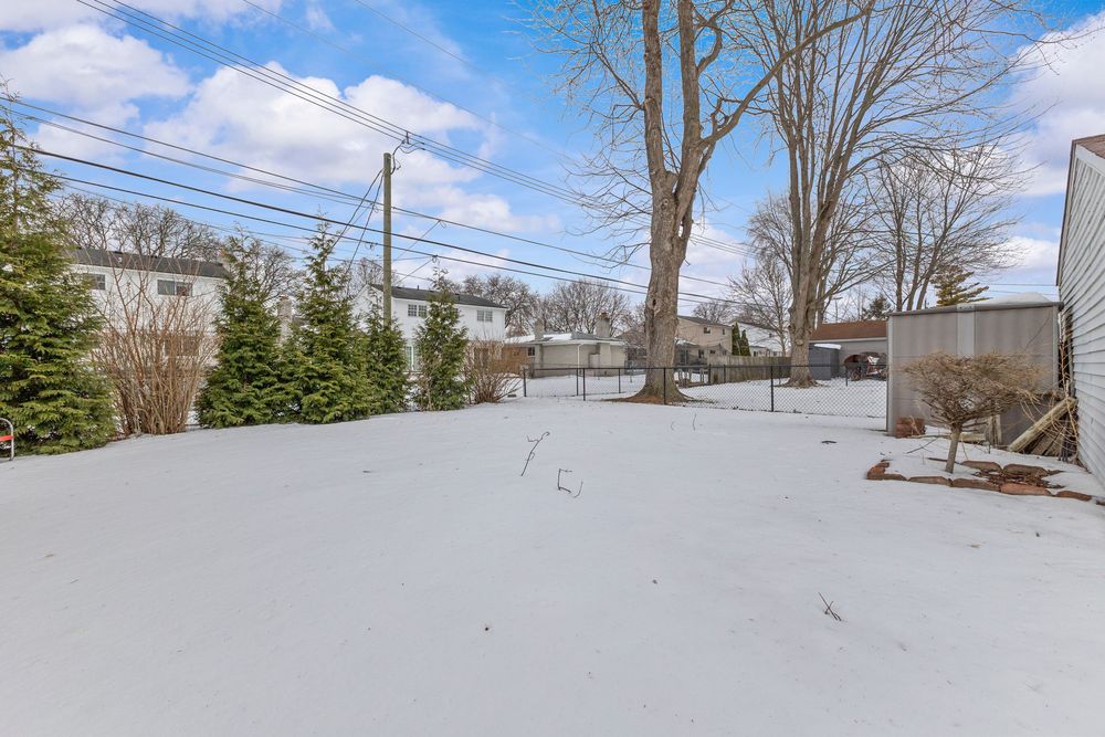 Snow-covered backyard with bare trees, evergreen shrubs, and a fence in a residential neighborhood under a partly cloudy sky.