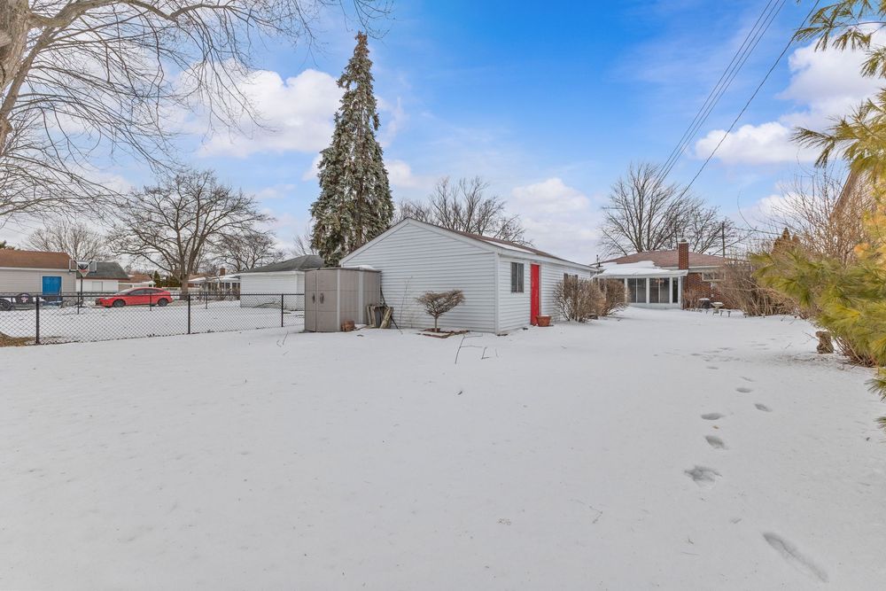 Snow-covered backyard with a white detached garage, red door accents, and bare trees under a partly cloudy winter sky.