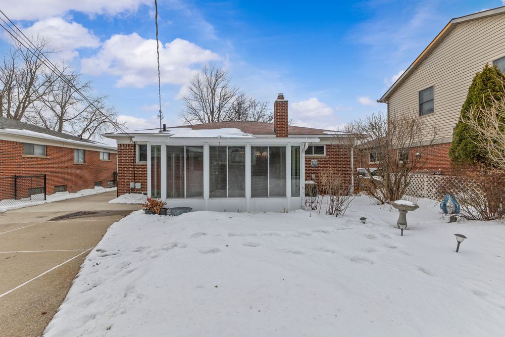 Snow-covered backyard of a brick ranch home with a sunroom addition and chimney under a partly cloudy winter sky.