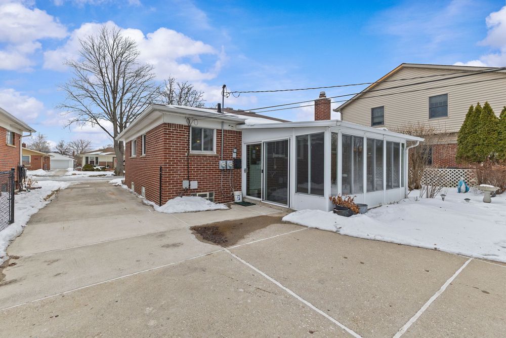 Brick ranch home with sunroom addition, snowy yard, and wide driveway under a partly cloudy winter sky.