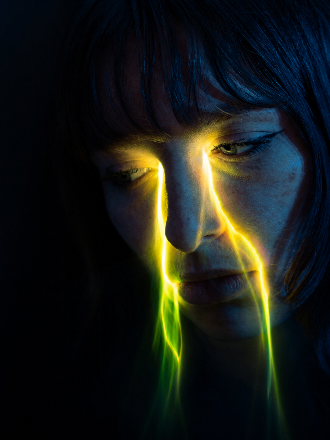 Woman with glowing yellow-green light streaks across her face in dramatic dark moody portrait photography.