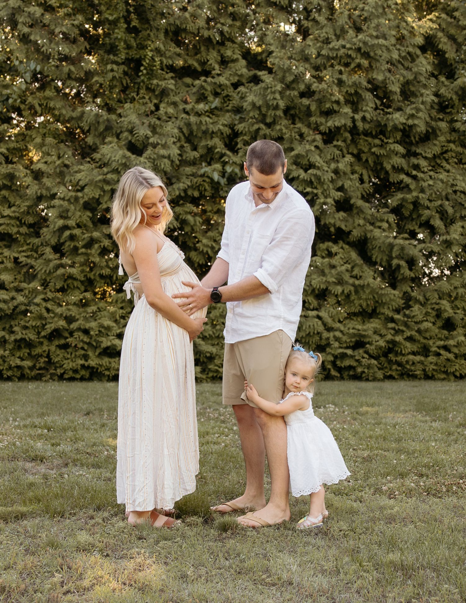 Pregnant woman in white dress with husband and toddler daughter posing outdoors in lush green garden.