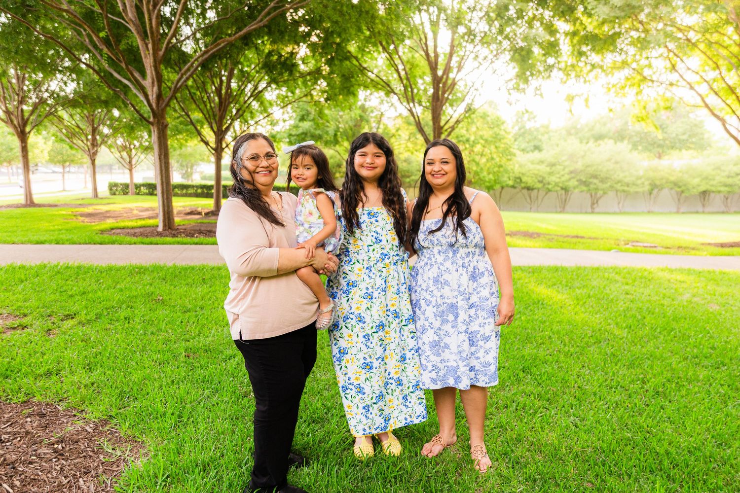 Four women smiling together outdoors in a lush green park surrounded by trees on a sunny day.
