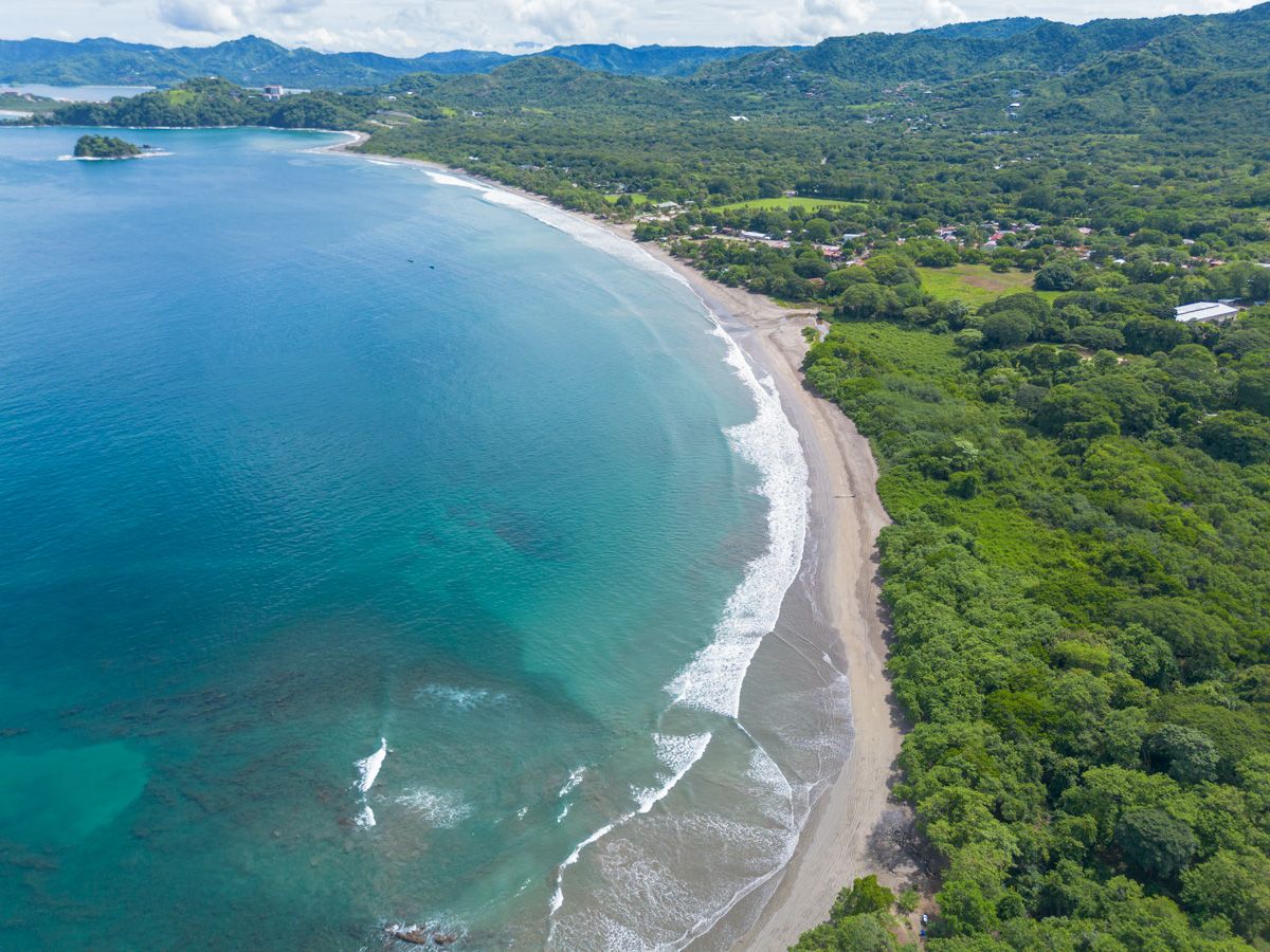 Aerial view of a curved tropical beach with turquoise waters, dark sand, and lush green jungle coastline.