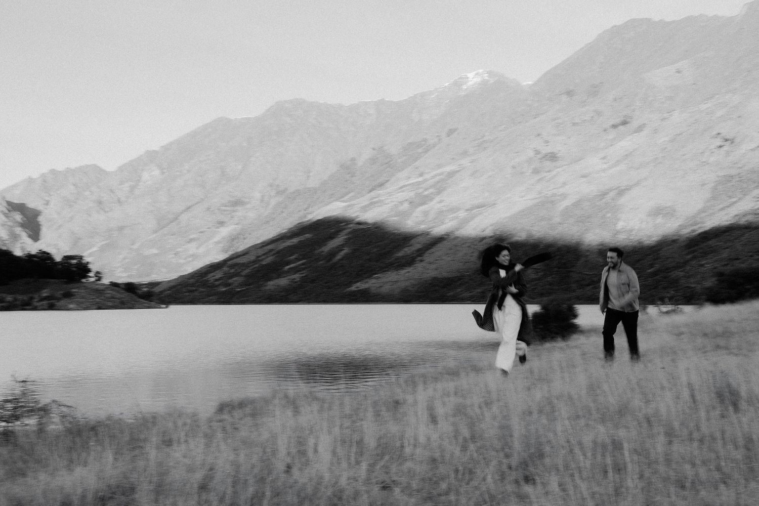 A couple walks along a lakeside meadow with dramatic mountain peaks in the background, captured in black and white.