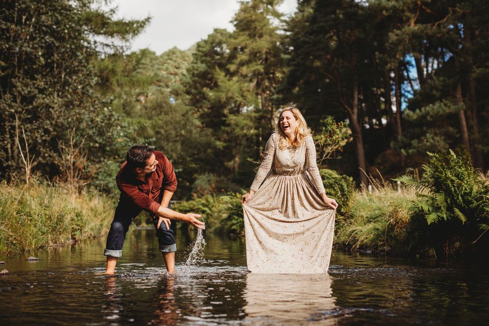 Couple laughing and playing in a shallow forest river, woman in elegant gold gown holding skirt up.